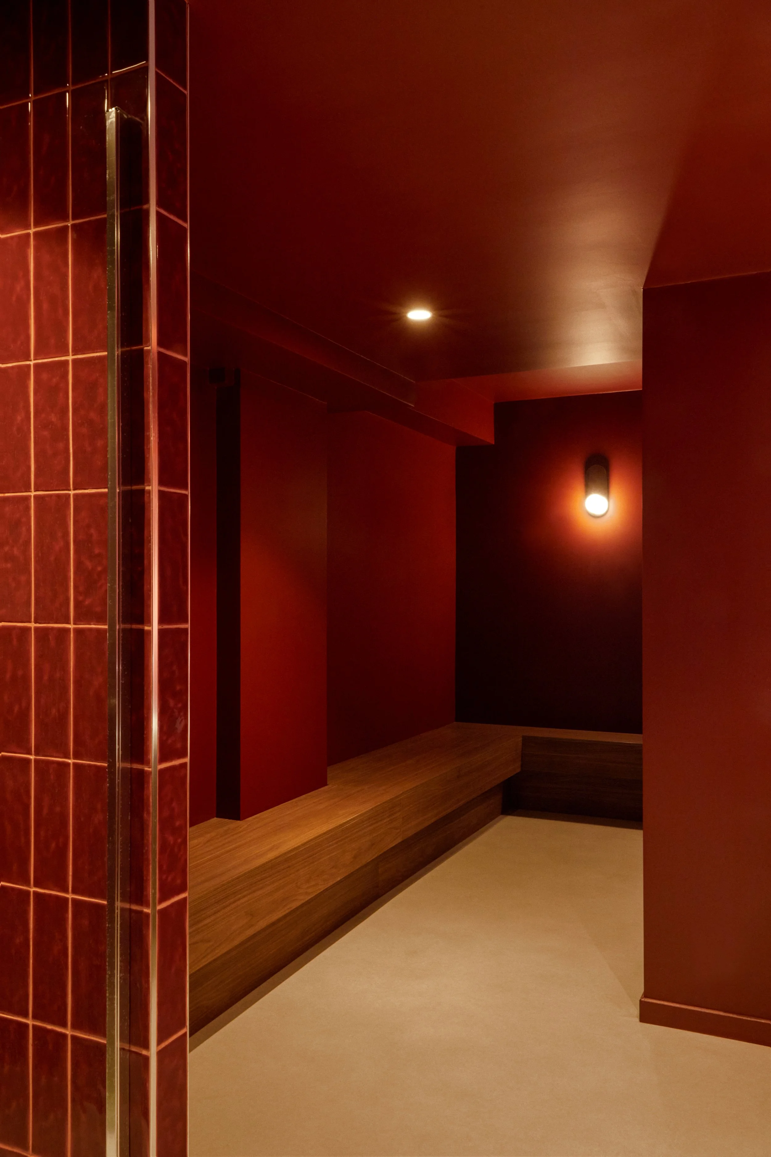 Aménagement d’un vestiaire et espace recovery pour studio wellness à Paris, avec banquette bois, murs brun-rouge et atmosphère enveloppante.