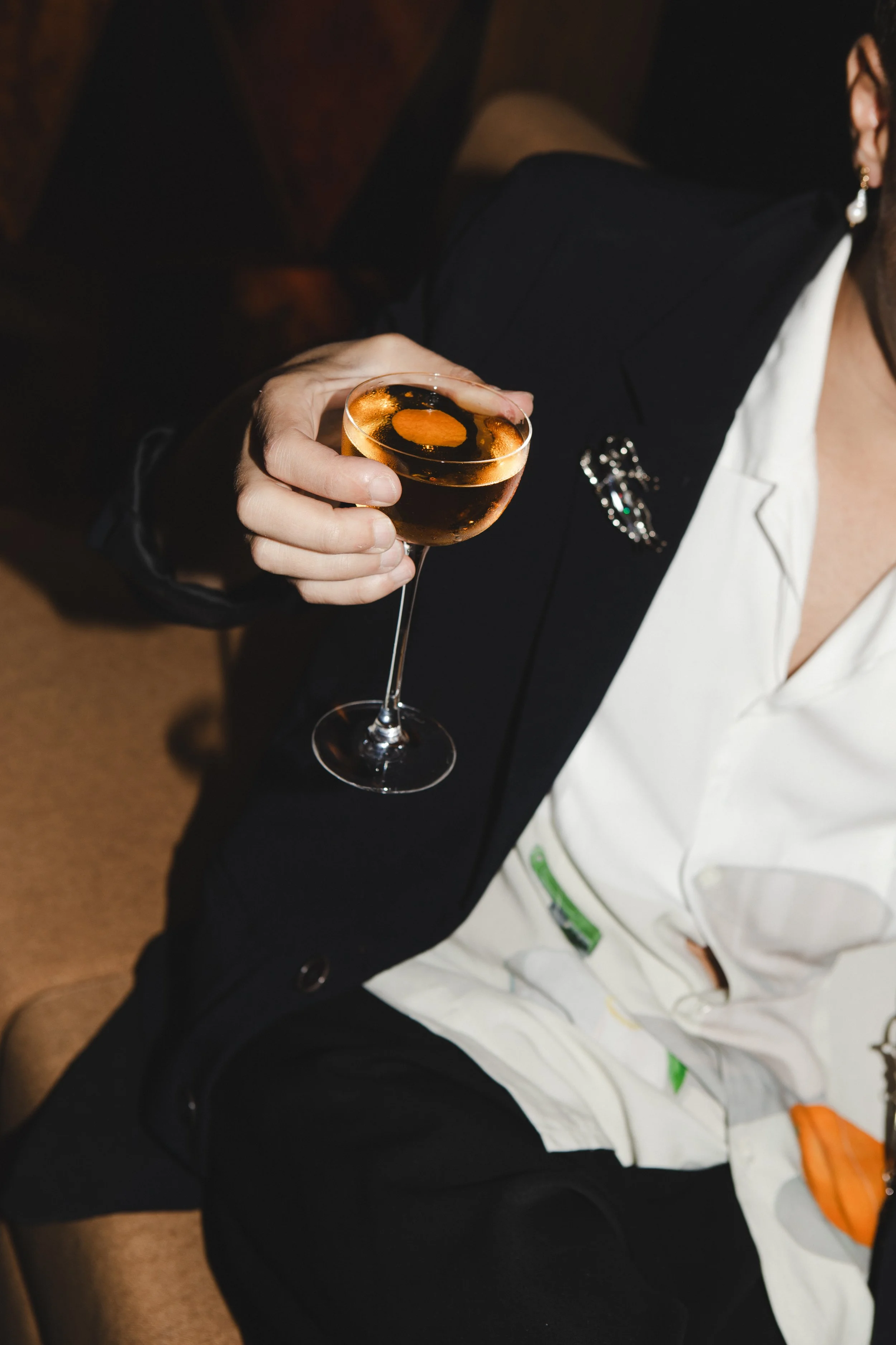 A person wearing black and white clothing holding a glass of dark-colored cocktail with ice in a dimly lit setting.
