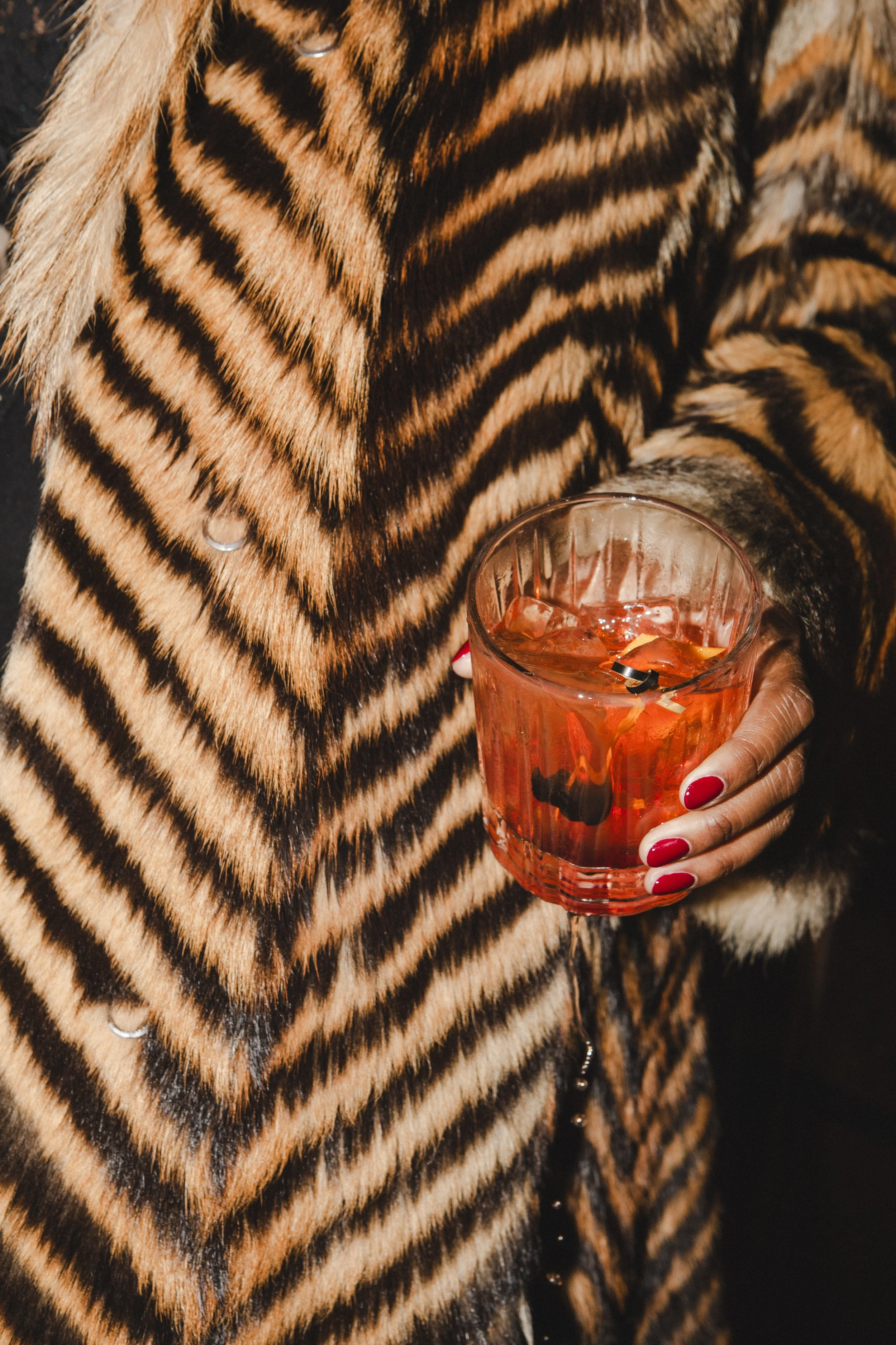 Person wearing a tiger-print fur coat holding a glass of cocktail with ice and a cherry.