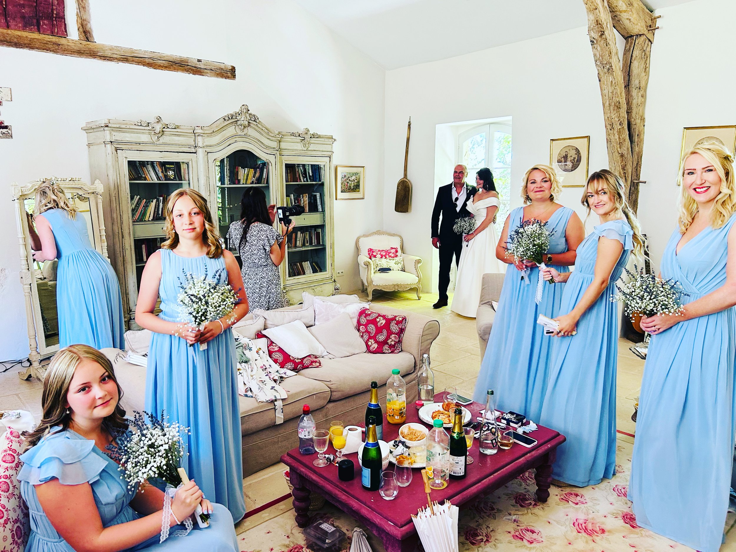 Bridal party and guests in a living room, some women in matching blue dresses holding small bouquets, a woman in white wedding dress, a man in a suit, guests drinking and socializing, cluttered table with drinks and snacks, old bookshelf and decorati