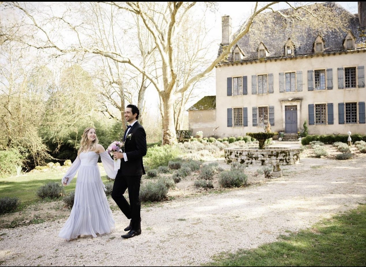 A bride and groom walking outdoors near a historic house, with the bride holding a bouquet of flowers, in a garden setting during the daytime.