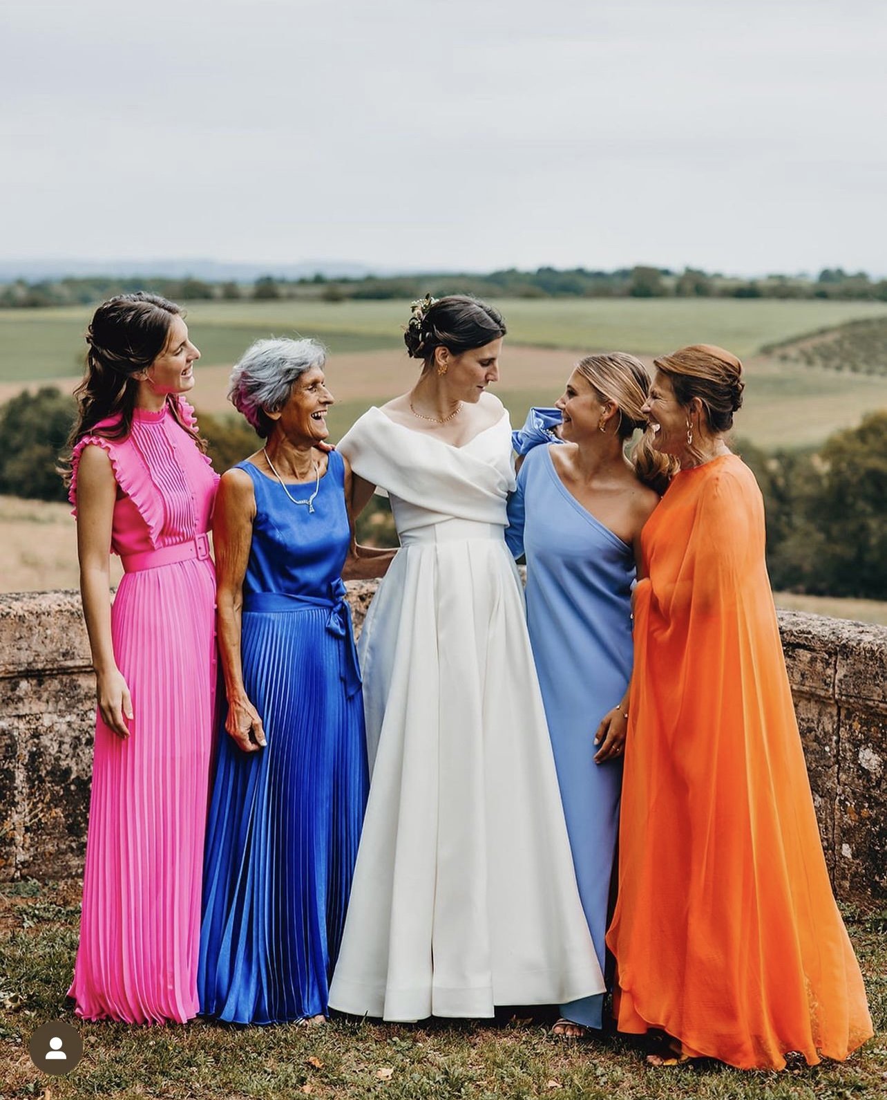 A group of women, including a bride in white, standing outdoors on a stone wall, dressed in colorful formal dresses, smiling and enjoying each other's company with a scenic landscape of fields and trees in the background.