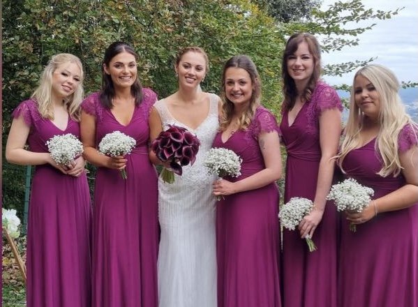 Bride in a white wedding gown standing with five bridesmaids in matching purple dresses, holding white bouquets, outdoors with trees in the background.