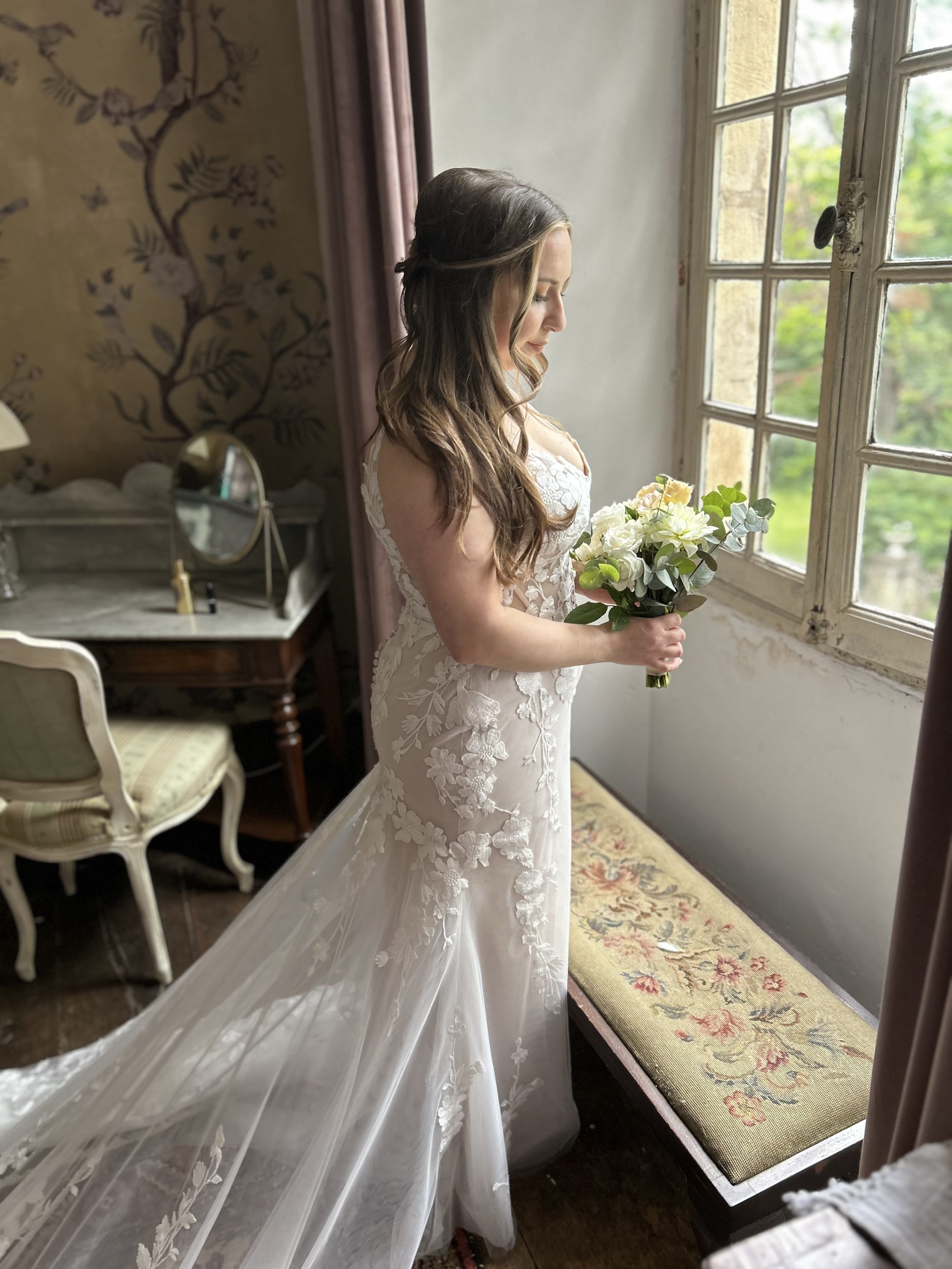 A bride in a wedding dress holding a bouquet, standing by an open window and looking down.