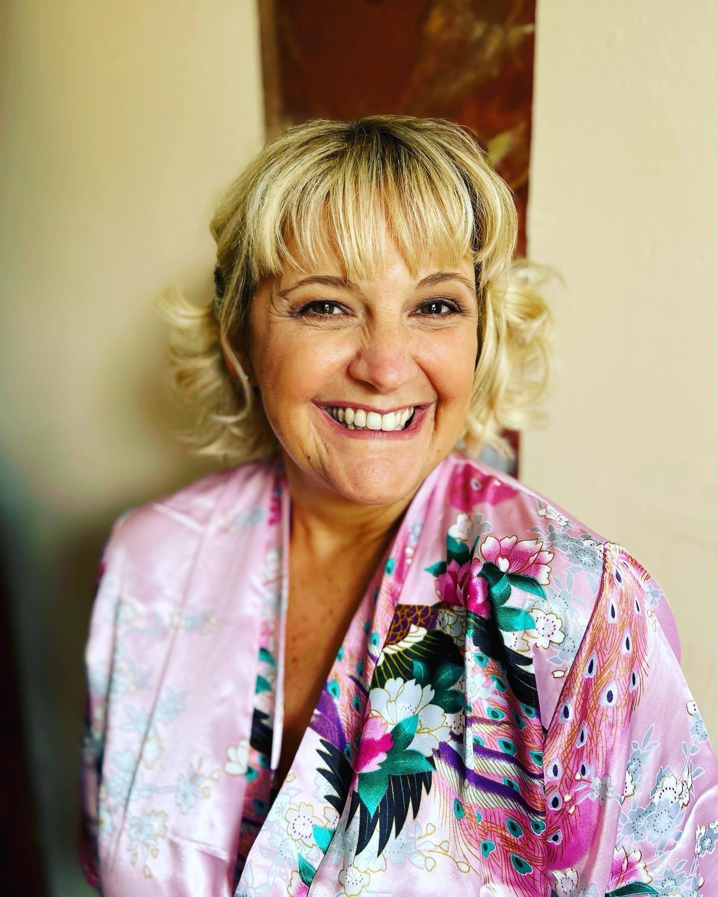 A smiling woman with blonde, curly hair wearing a pink floral kimono. She is standing indoors against a beige wall with part of an abstract painting behind her.