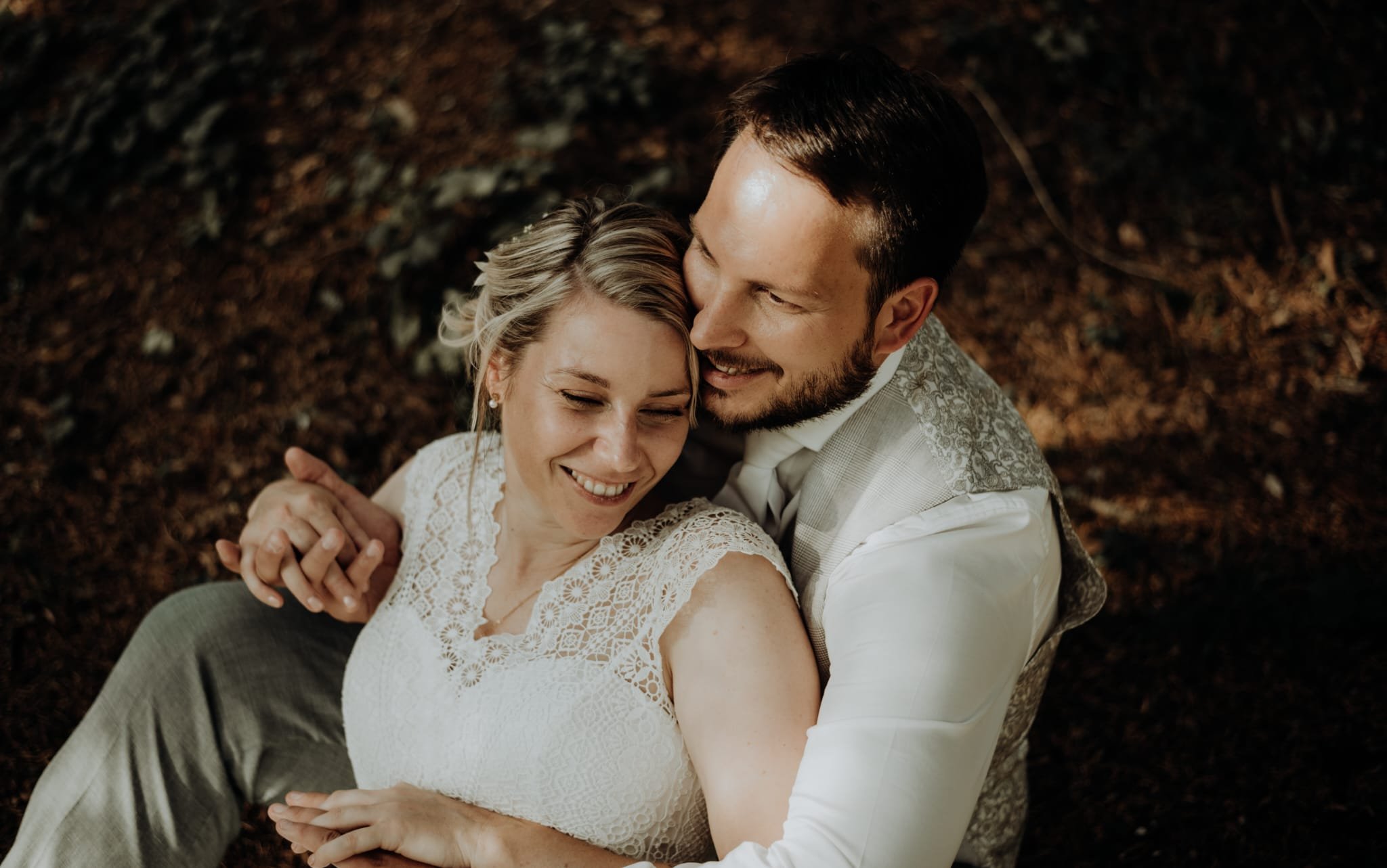 A happy couple sitting on the ground outdoors, embracing and smiling. The woman has blonde hair and is wearing a white lace dress, while the man has dark hair and is wearing a light-colored vest and shirt.