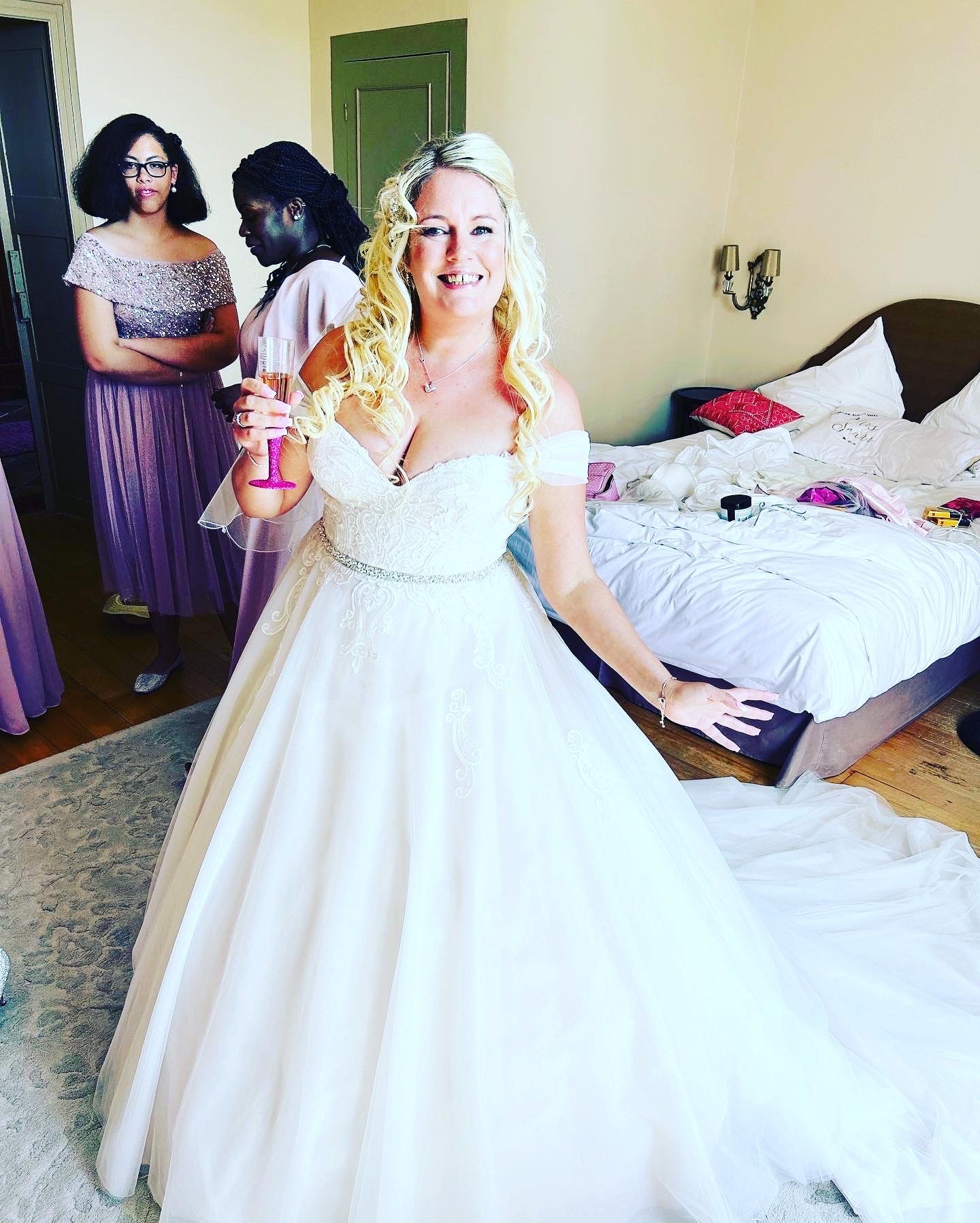 A bride in a white wedding dress holding a glass, smiling in a room with three women in purple dresses behind her and an unmade bed in the background.