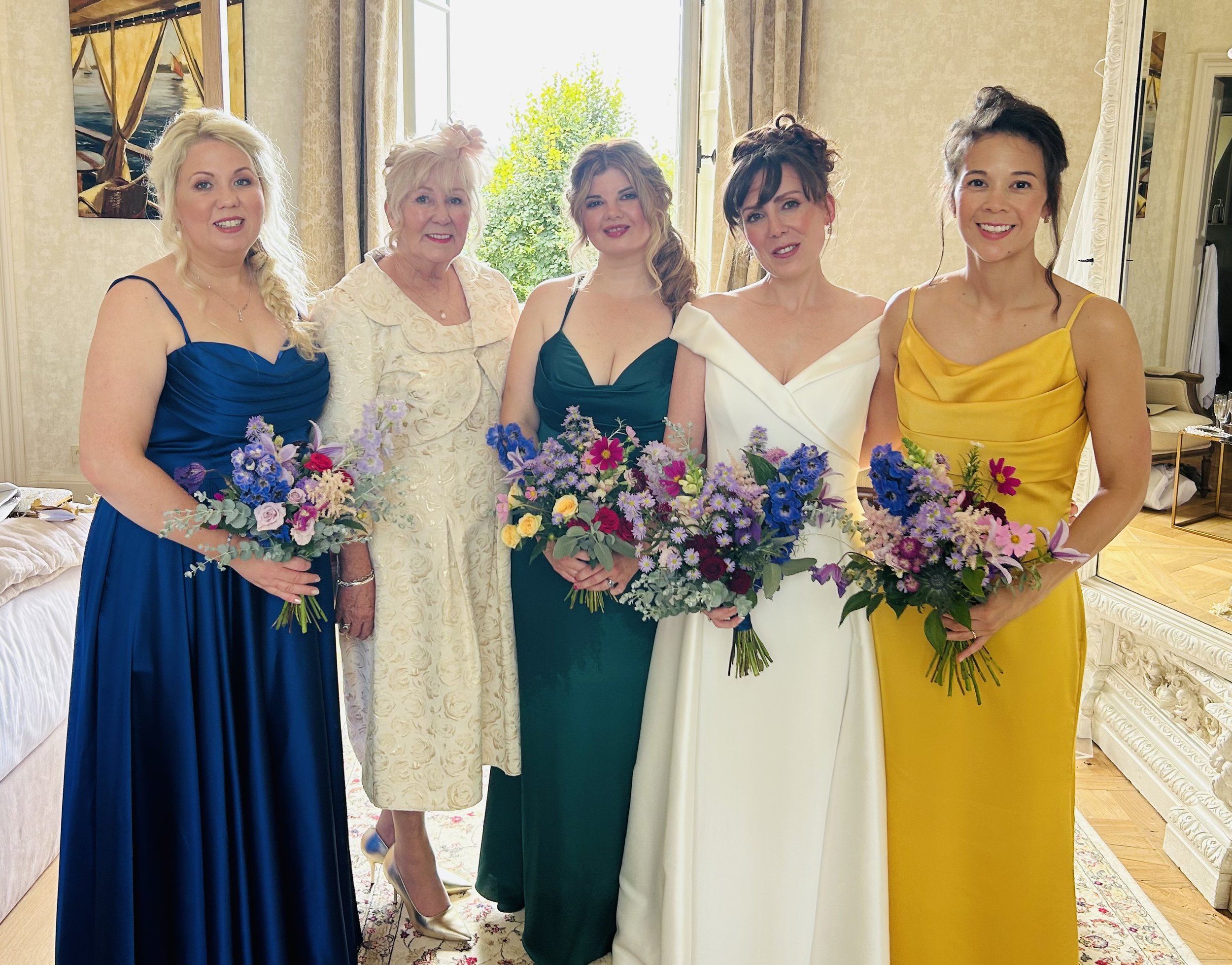 Five women in colorful dresses holding bouquets of flowers standing inside a room with large windows and artwork on the wall.
