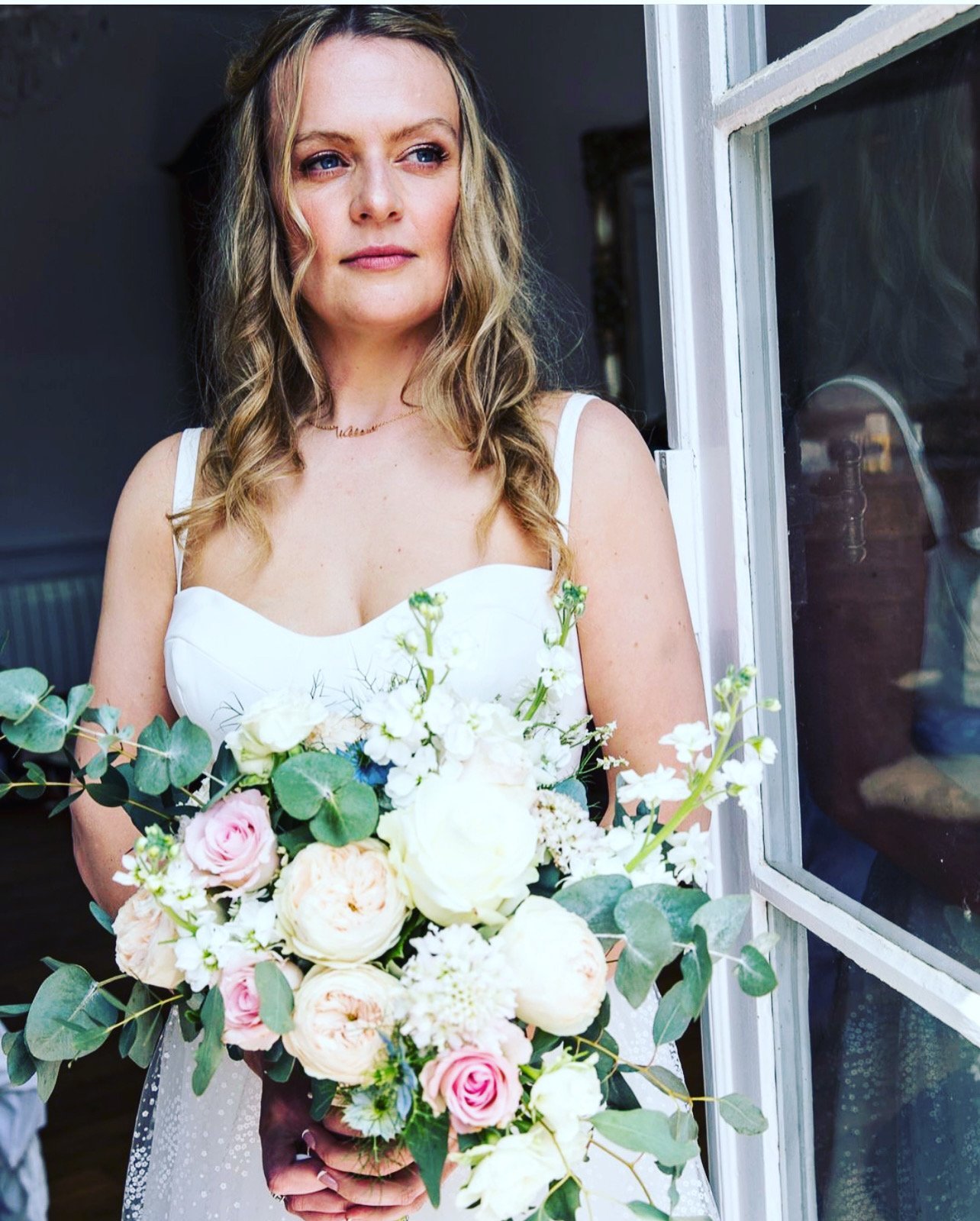 A bride with wavy blonde hair in a white dress holding a bouquet of flowers standing near a window.