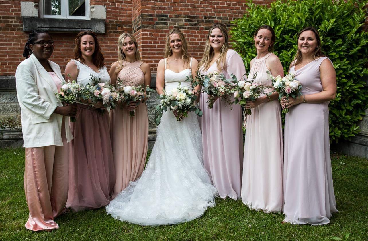 Bride in a white wedding gown surrounded by six bridesmaids in pastel-colored dresses holding bouquets, standing on grass in front of a brick building and green shrubbery.