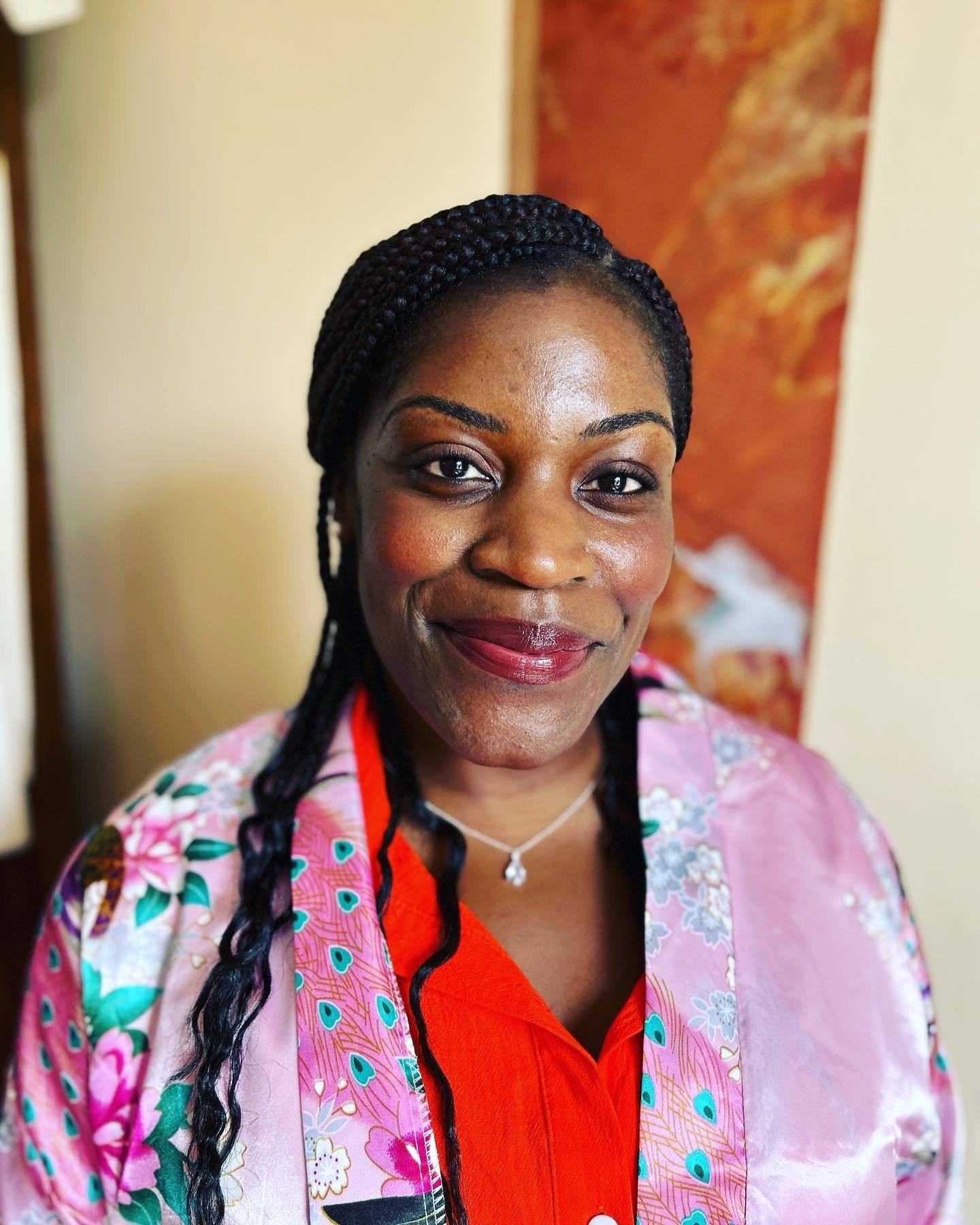 Close-up of a smiling woman with braided black hair wearing a pink floral robe over an orange top, standing indoors with a red and beige wall behind her.