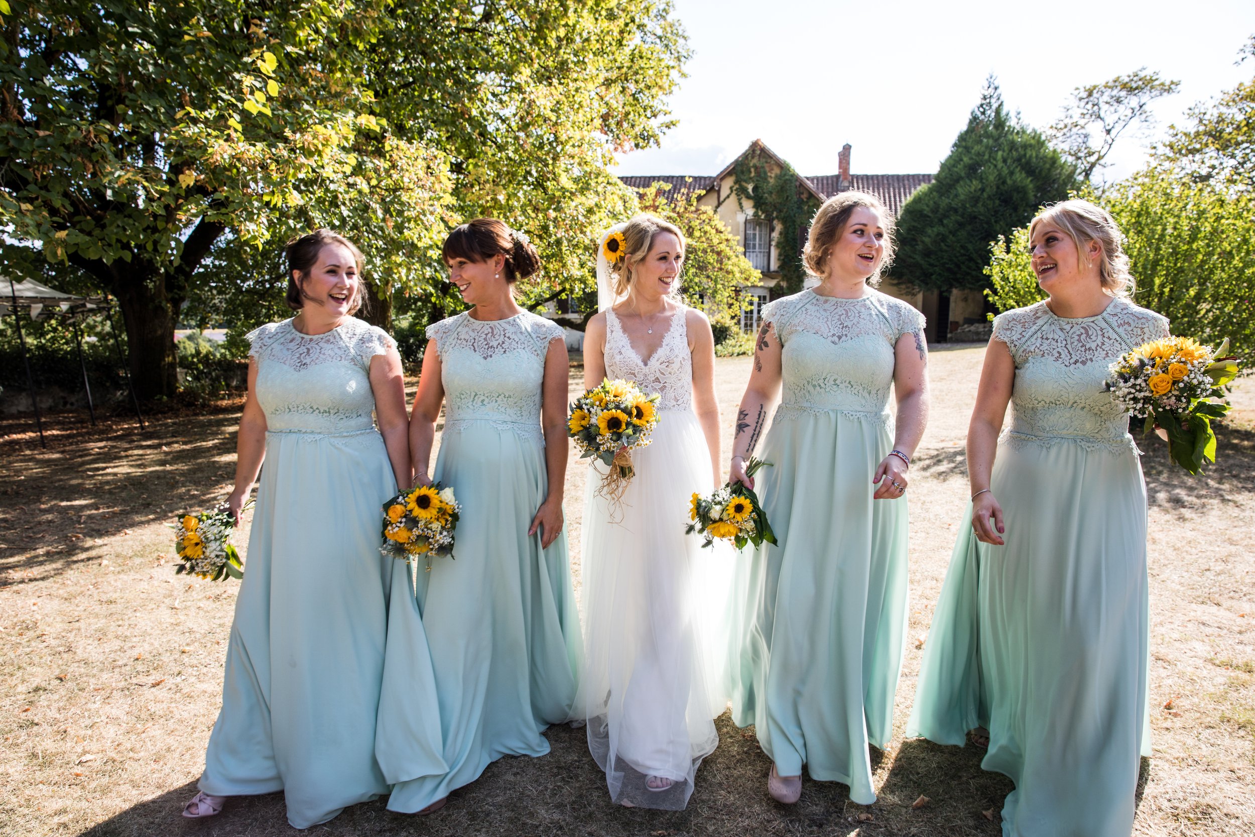 Bride and five bridesmaids walking outdoors on a sunny day, holding bouquets of sunflowers and white flowers, wearing light blue dresses with lace tops.