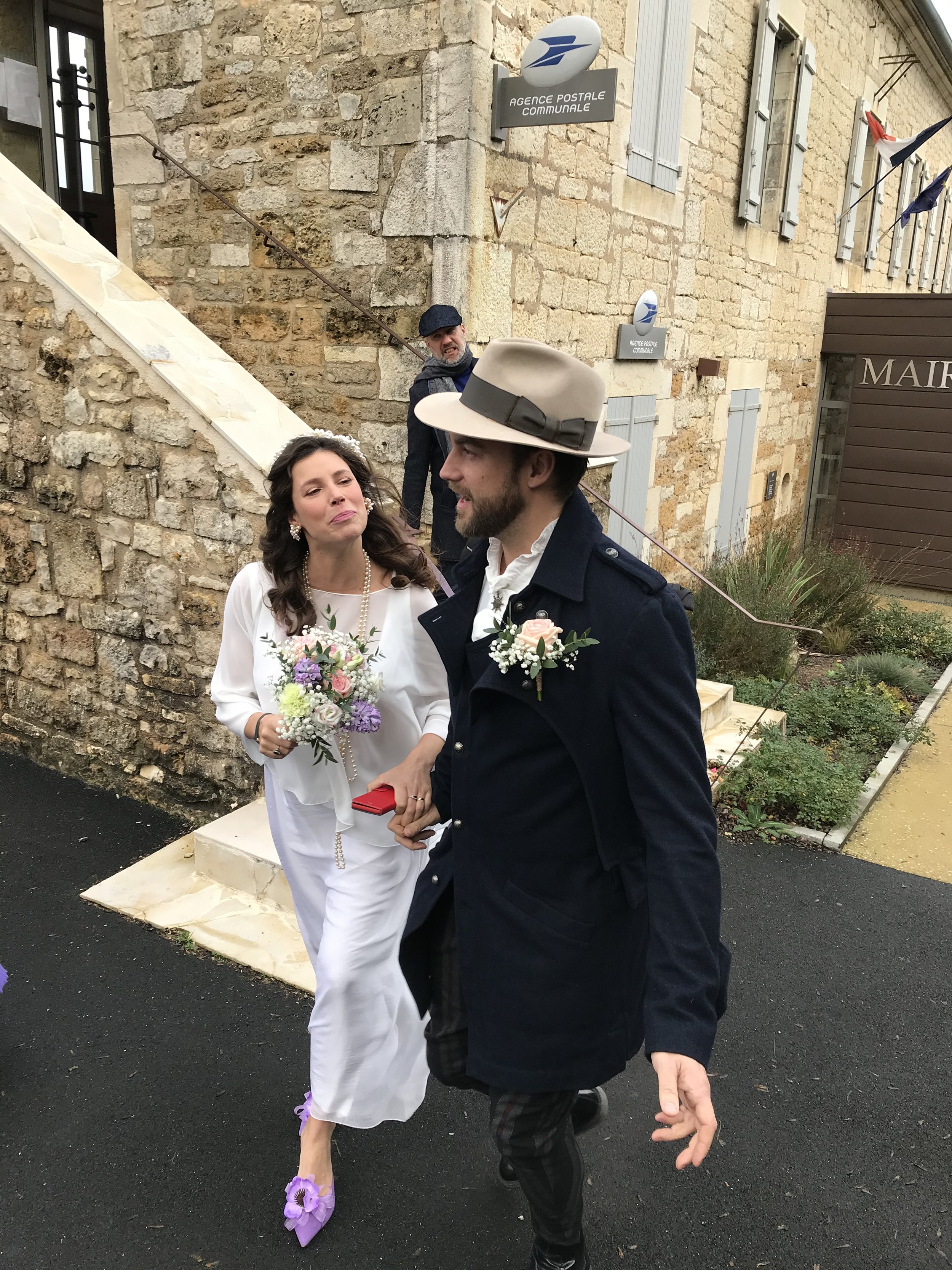 A bride and groom dressed in vintage-style wedding attire holding hands outside a stone building, with a man in the background watching. The bride is wearing a white dress, purple shoes, and holding a bouquet. The groom is wearing a dark coat, a hat,