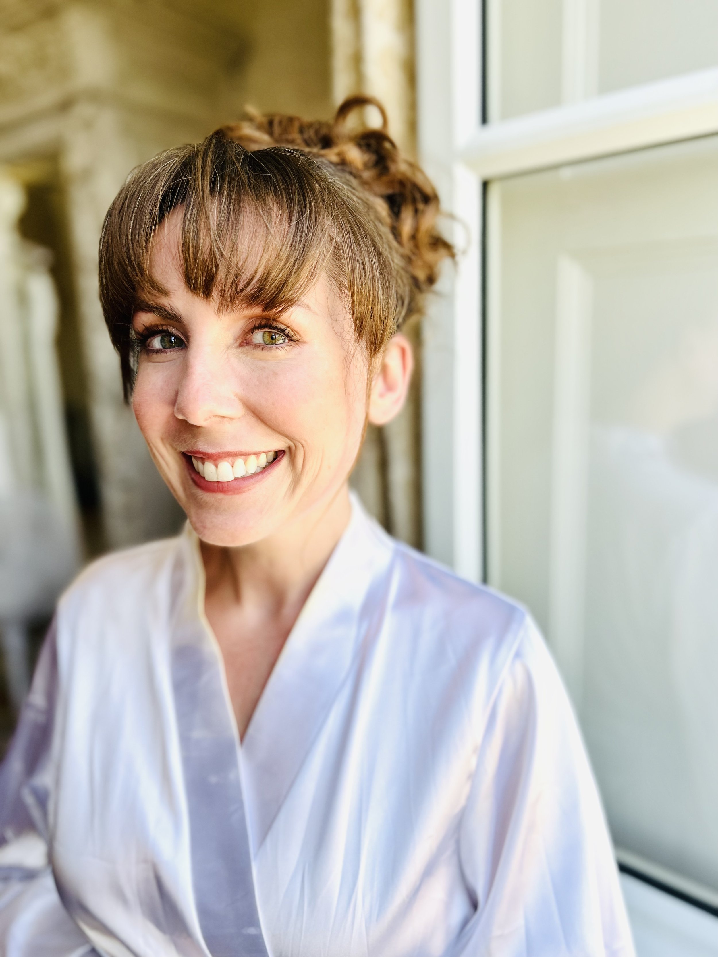A smiling woman with short brown hair and green eyes, wearing a white satin robe, sitting near a window with sunlight shining on her face.