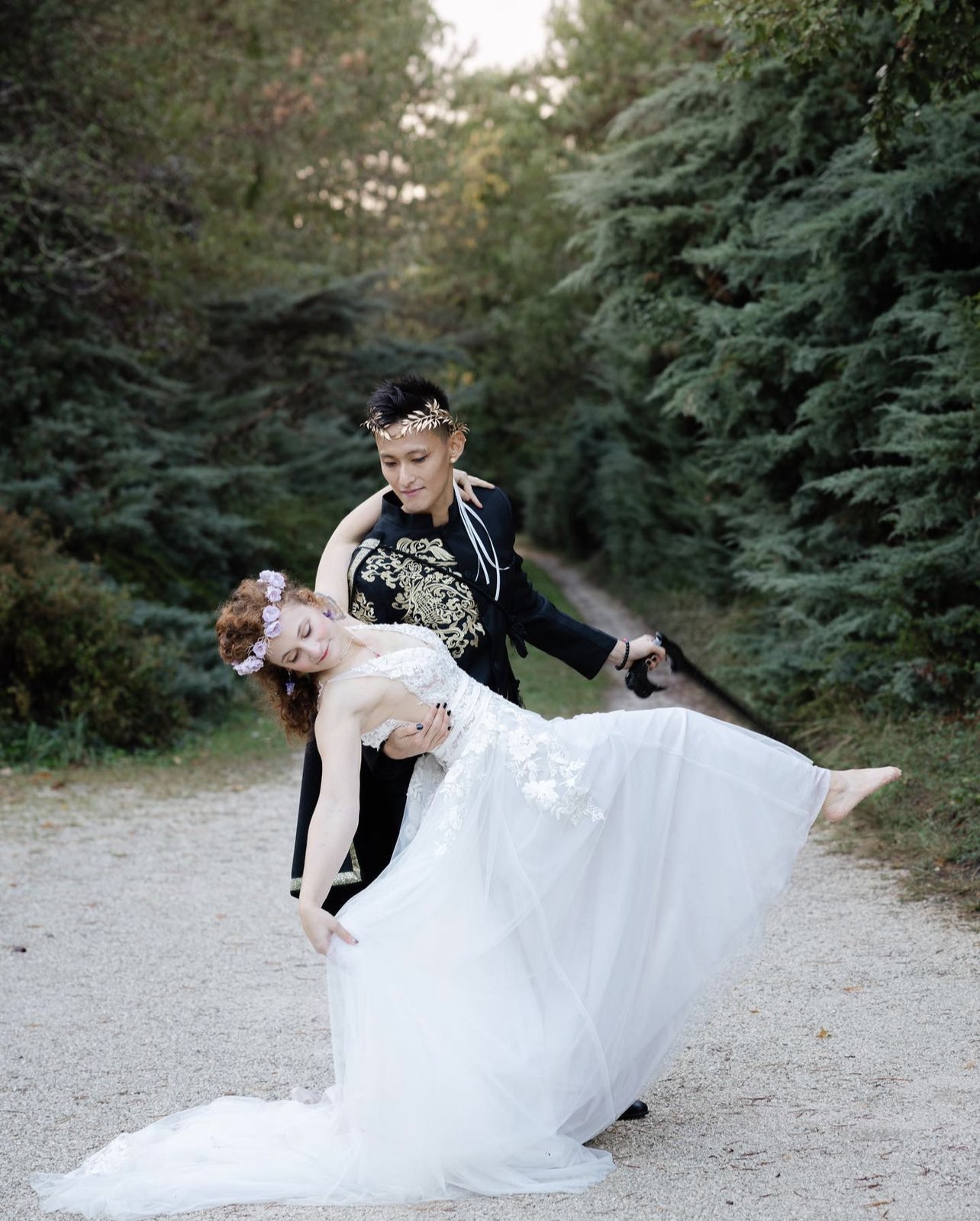 A bride and groom dancing outdoors on a nature trail surrounded by trees, with the groom holding the bride as she leans back with her eyes closed.
