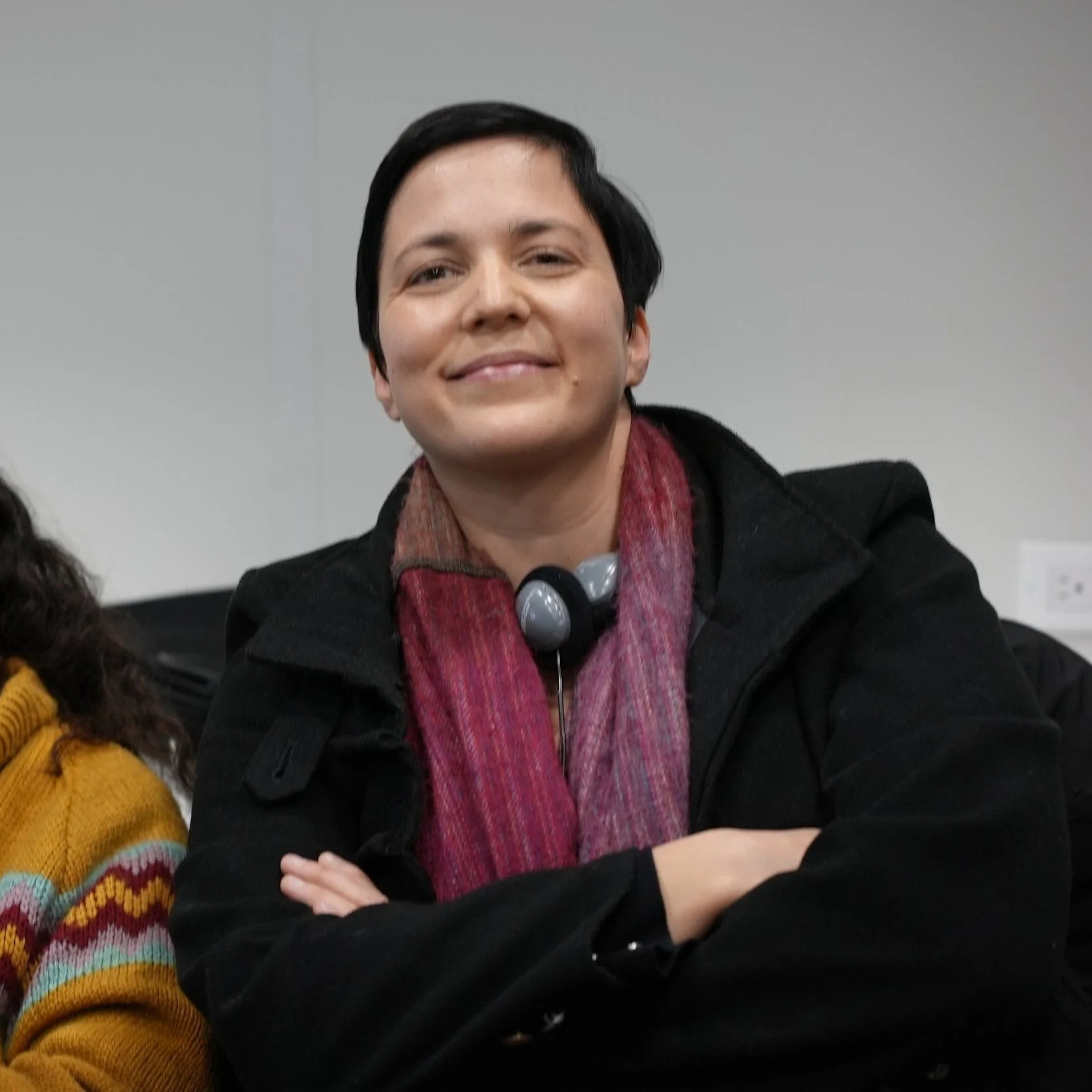 Smiling person with short black hair, wearing a black jacket and a pinkish-red scarf, sitting with arms crossed, in an indoor setting.