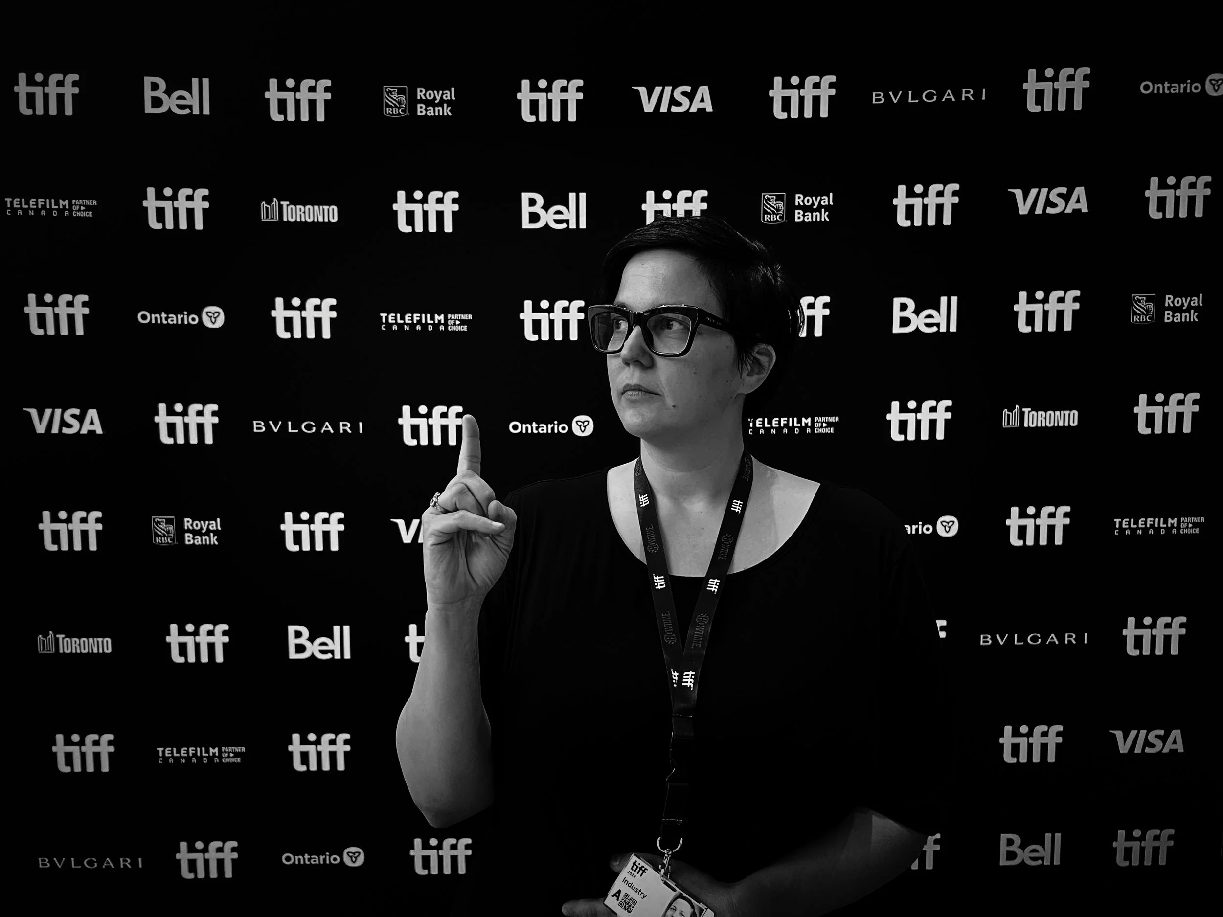 A woman with short dark hair and glasses stands in front of a backdrop with TIFF logos, wearing a lanyard and making a gesture with her index finger raised.