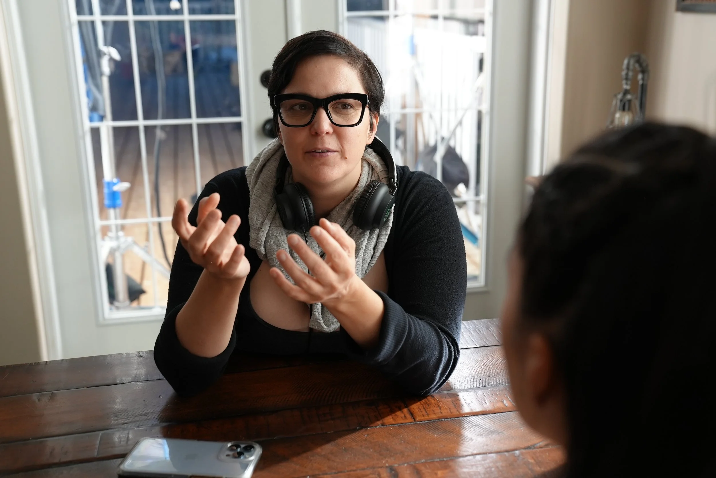 Woman with short dark hair, black glasses, and headphones around her neck, talking to another person with dark hair at a wooden table in a room with glass doors.