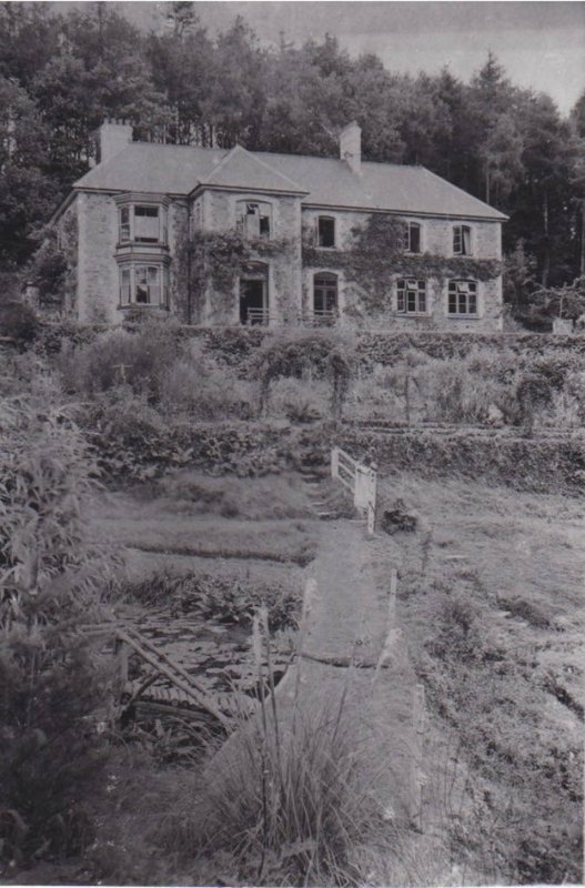 Black and white photo of an old two-story stone house with ivy-covered walls, surrounded by a garden and dense trees. In the foreground, a wooden footbridge over a small pond leads toward the house.