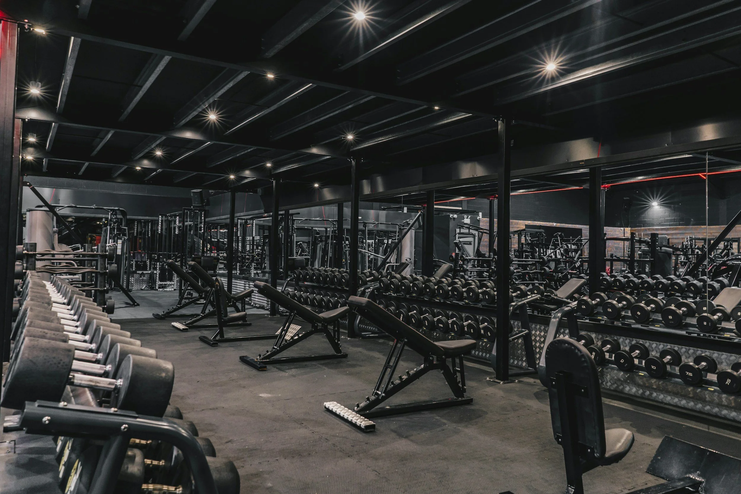 Interior of a gym with rows of dumbbells, equipment, and workout benches, illuminated by ceiling lights.