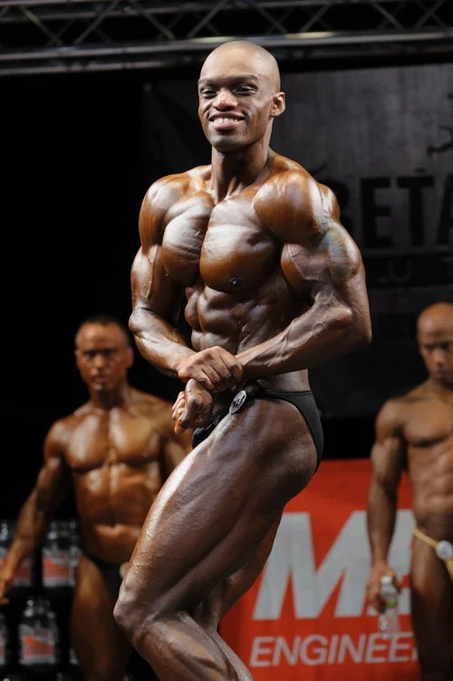 A male bodybuilder with dark skin and a shaved head flexes his muscles on stage during a bodybuilding competition, with two other shirtless bodybuilders in the background.