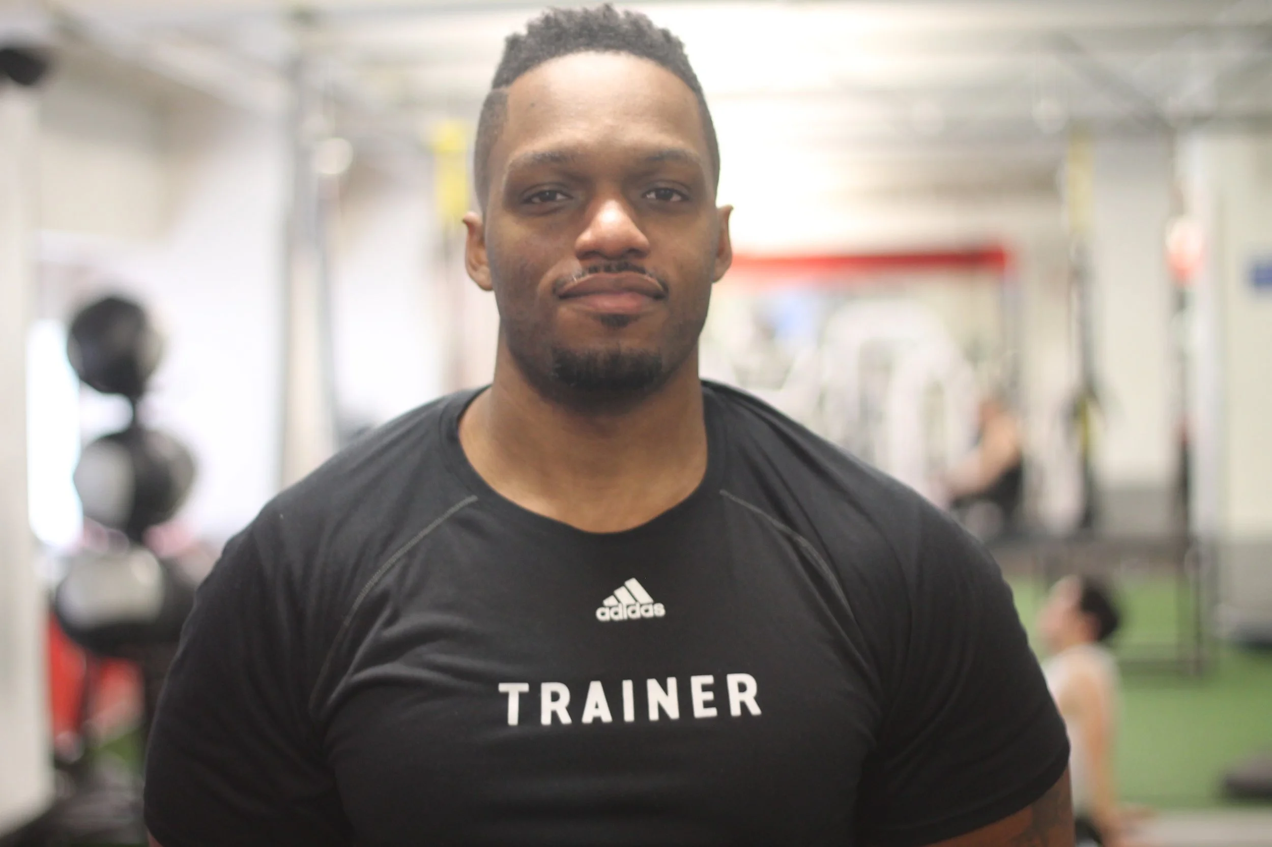 A man with short hair and a goatee smiling slightly, wearing a black athletic shirt with 'TRAINER' written on it, standing in a gym with workout equipment in the background.