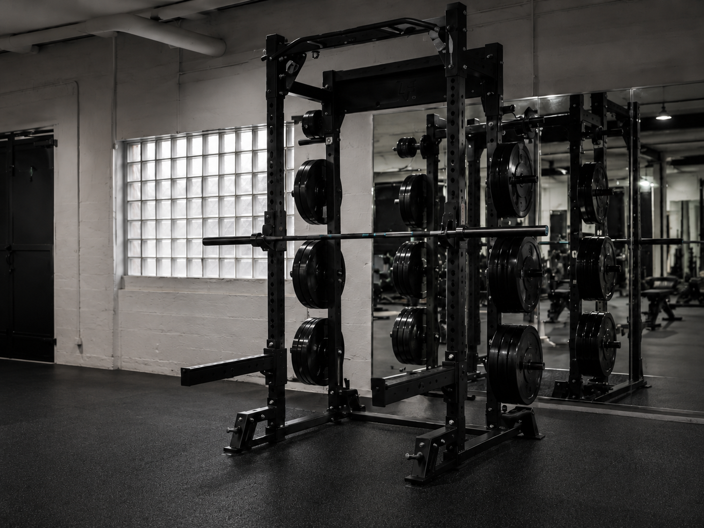 Empty gym with black metal squat rack holding weight plates and a barbell, mirror reflecting gym equipment, concrete wall with glass block window, and rubber gym floor.