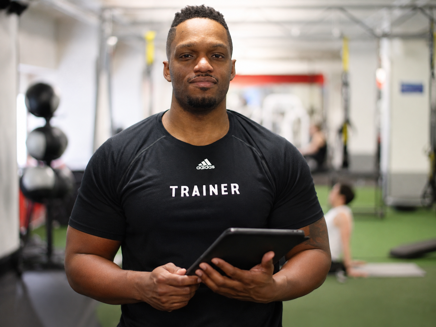 A male fitness trainer wearing a black 'TRAINER' shirt holding a tablet inside a gym, with gym equipment and a person doing yoga in the background.