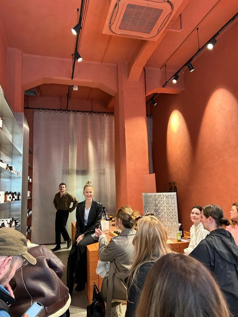 Group of people sitting and listening to a woman speaking in an indoor venue with warm orange walls, shelves with bottles, and a gray curtain in the background.