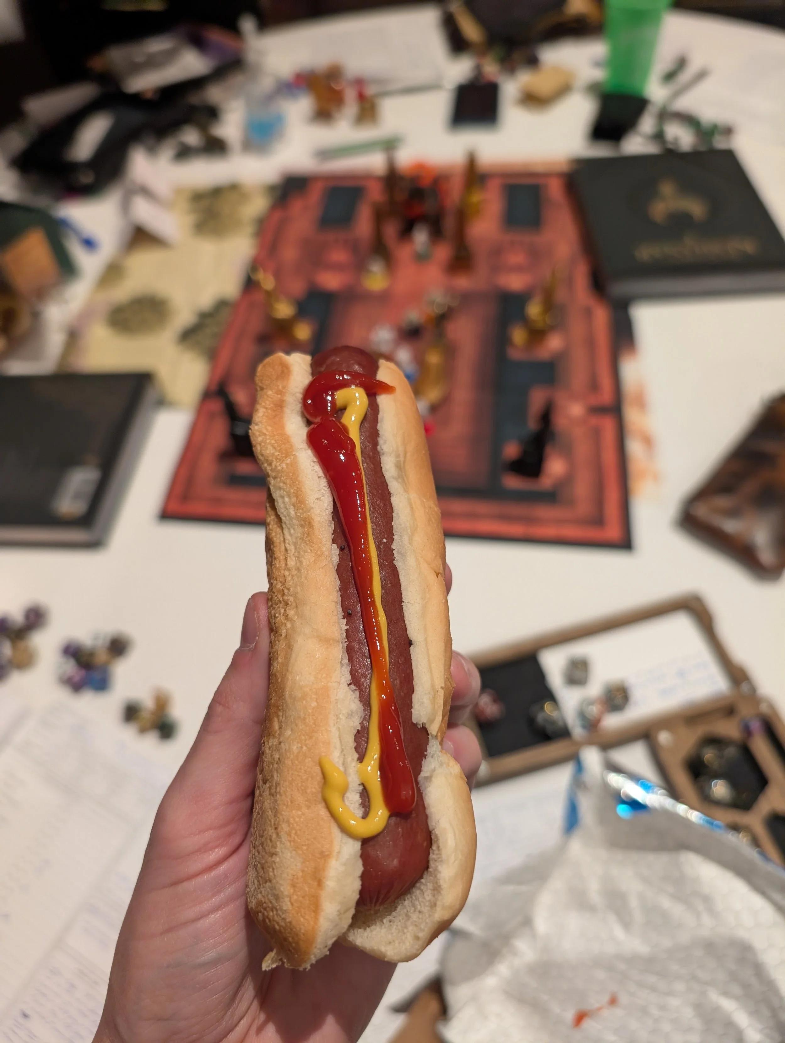 Susan holding a hot dog with ketchup and mustard over a crowded gaming table.