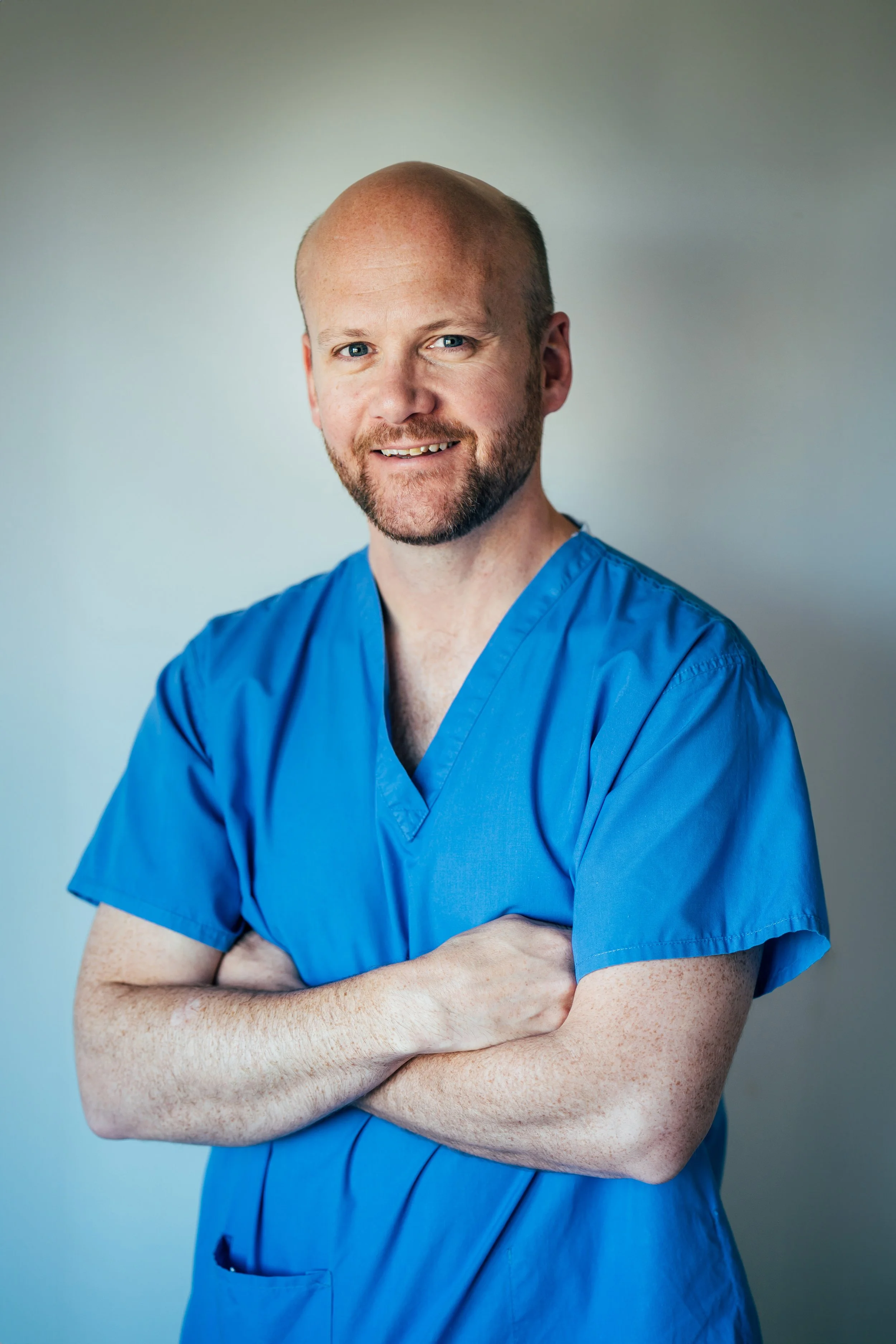 A man with a bald head and beard wearing blue medical scrubs, smiling with arms crossed, standing against a neutral background.