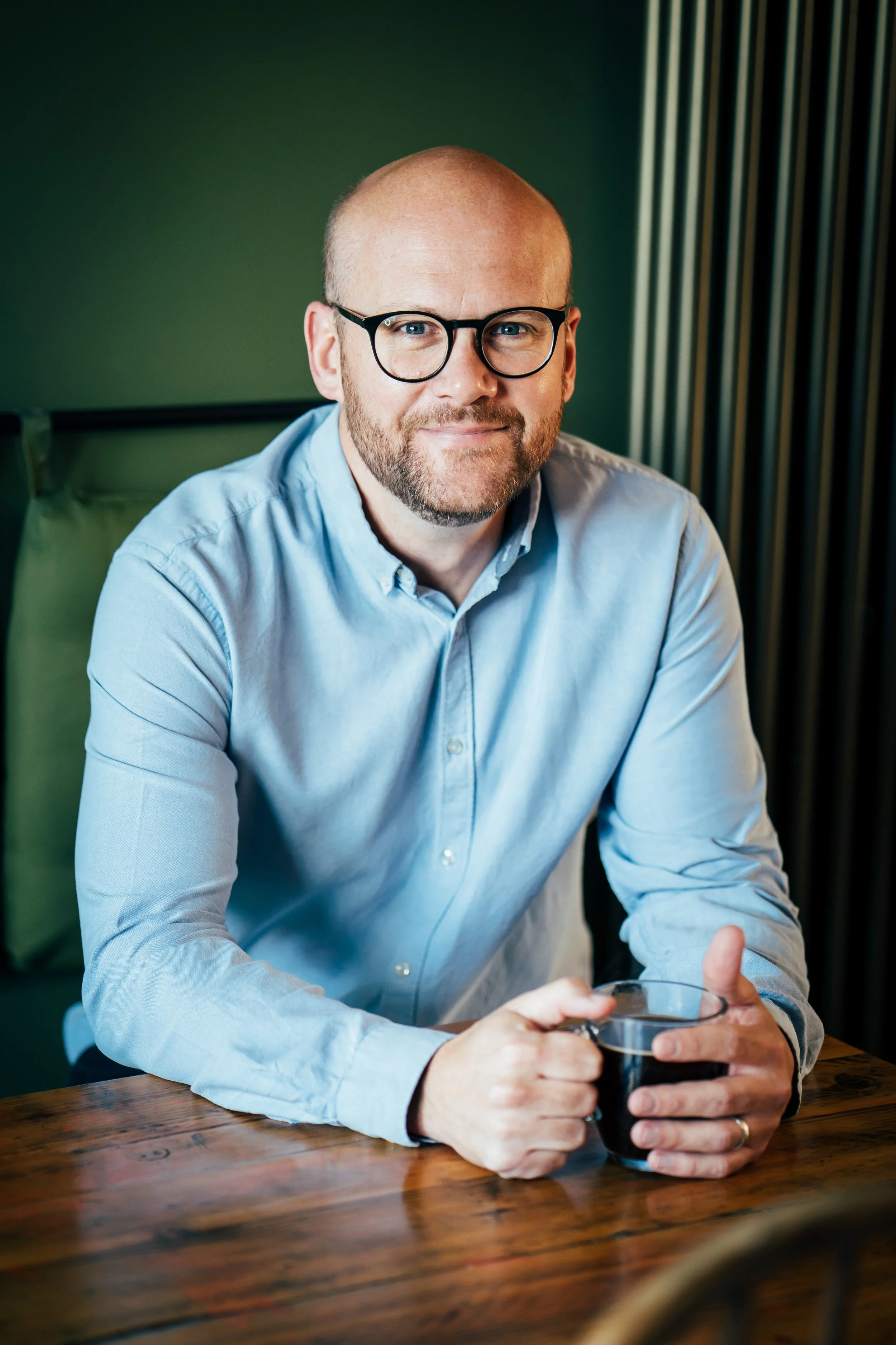 A man with glasses, a bald head, and a beard, wearing a light blue button-up shirt, sitting at a wooden table with a dark beverage in a clear mug.