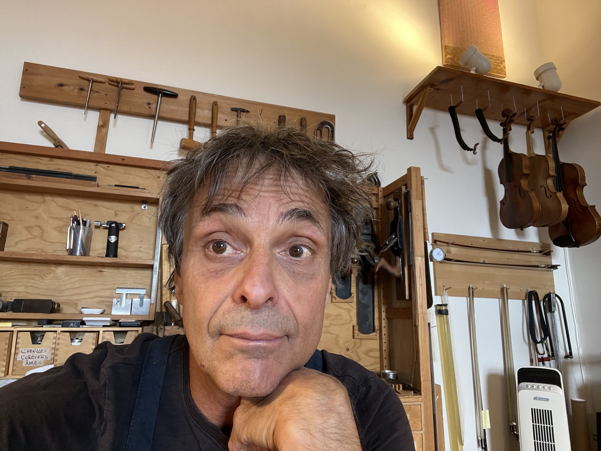 A man resting his chin on his hand in a woodworking workshop with various hand tools and stringed instruments hanging on the wall behind him.