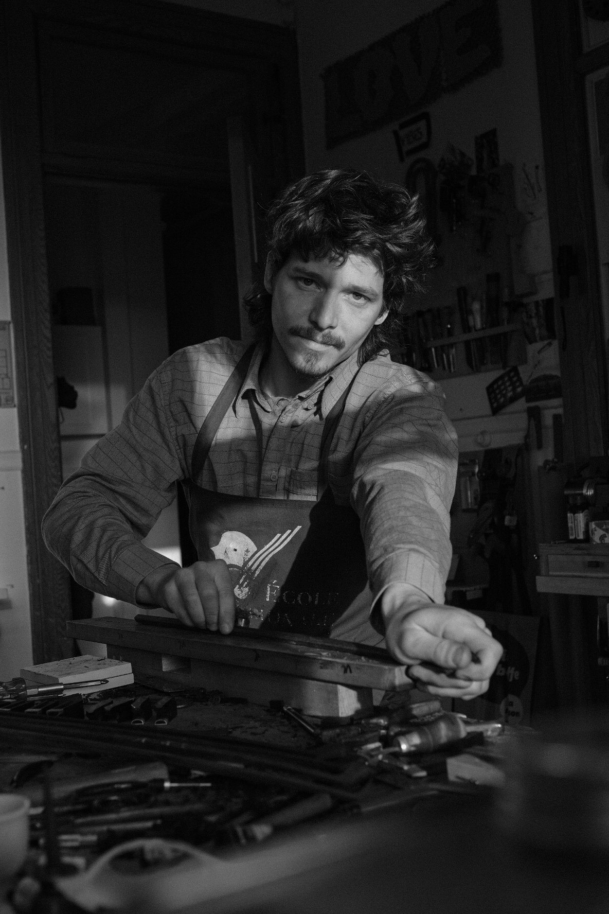 A man with wavy hair and a mustache, wearing an apron, is working in a workshop surrounded by tools and equipment, reaching out towards the camera.