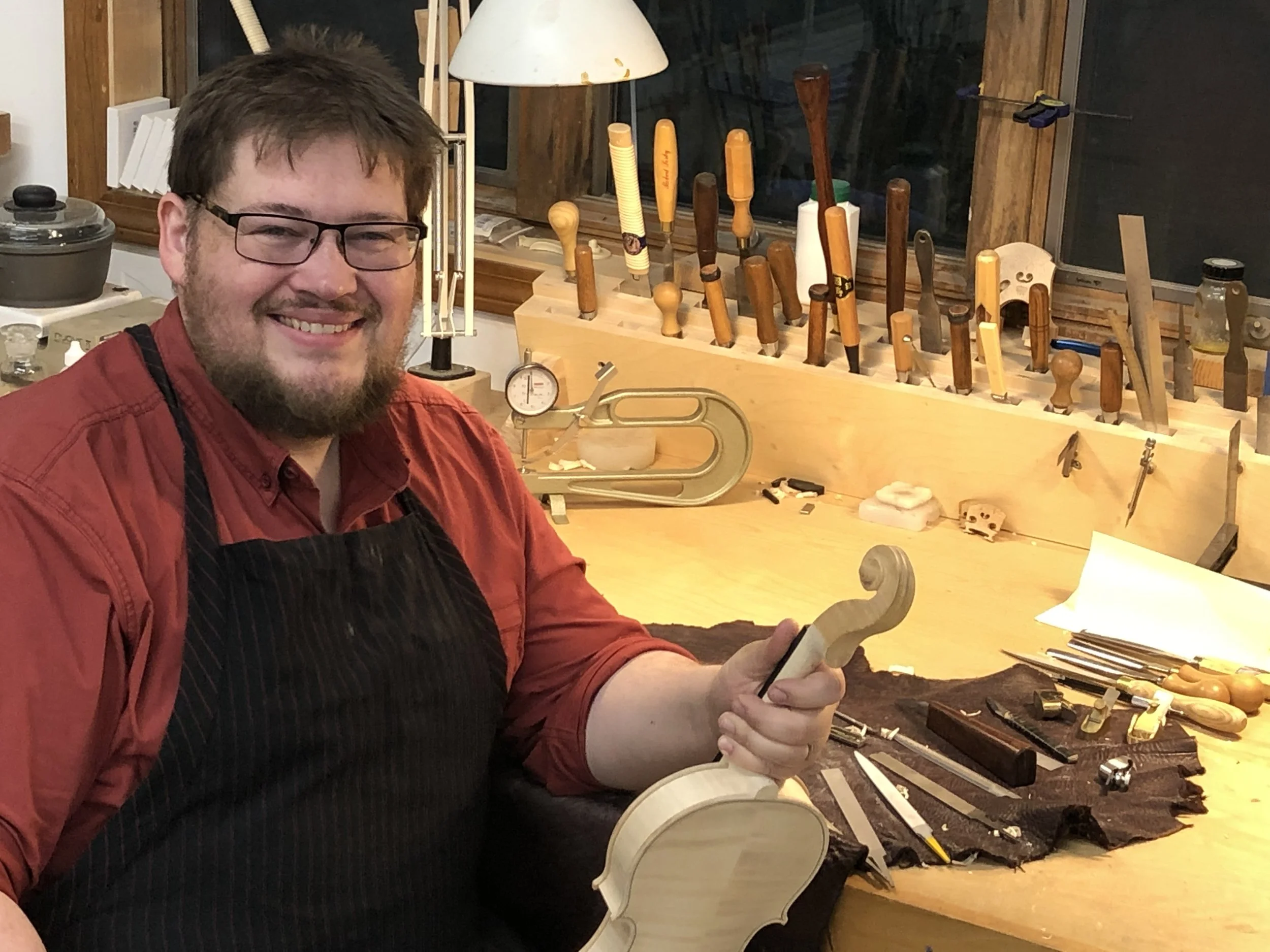 A smiling man with glasses and a beard sitting at a woodworking bench, holding a carved violin scroll in his hand, surrounded by woodworking tools and a work-in-progress violin.