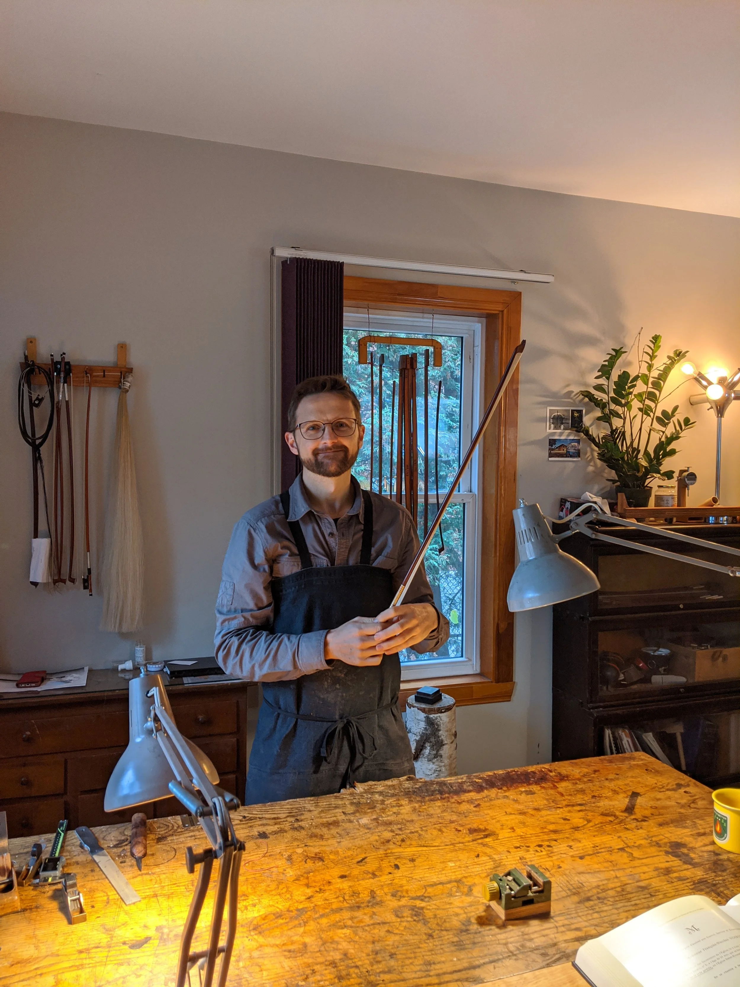 A man standing behind a large wooden worktable holding a glass cutter tool in a workshop. He is wearing glasses and a black apron, with various tools and supplies on the table. The background includes a window, a plant, and a lamp.