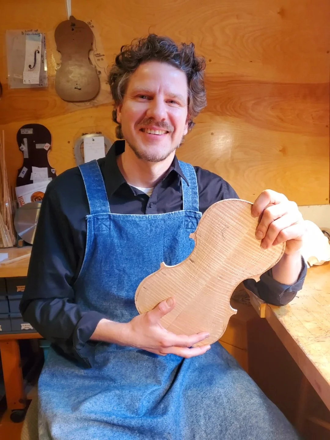 A man with curly hair and a beard smiling and holding a piece of unfinished wood shaped like a musical instrument, possibly a violin, in a woodworking shop.