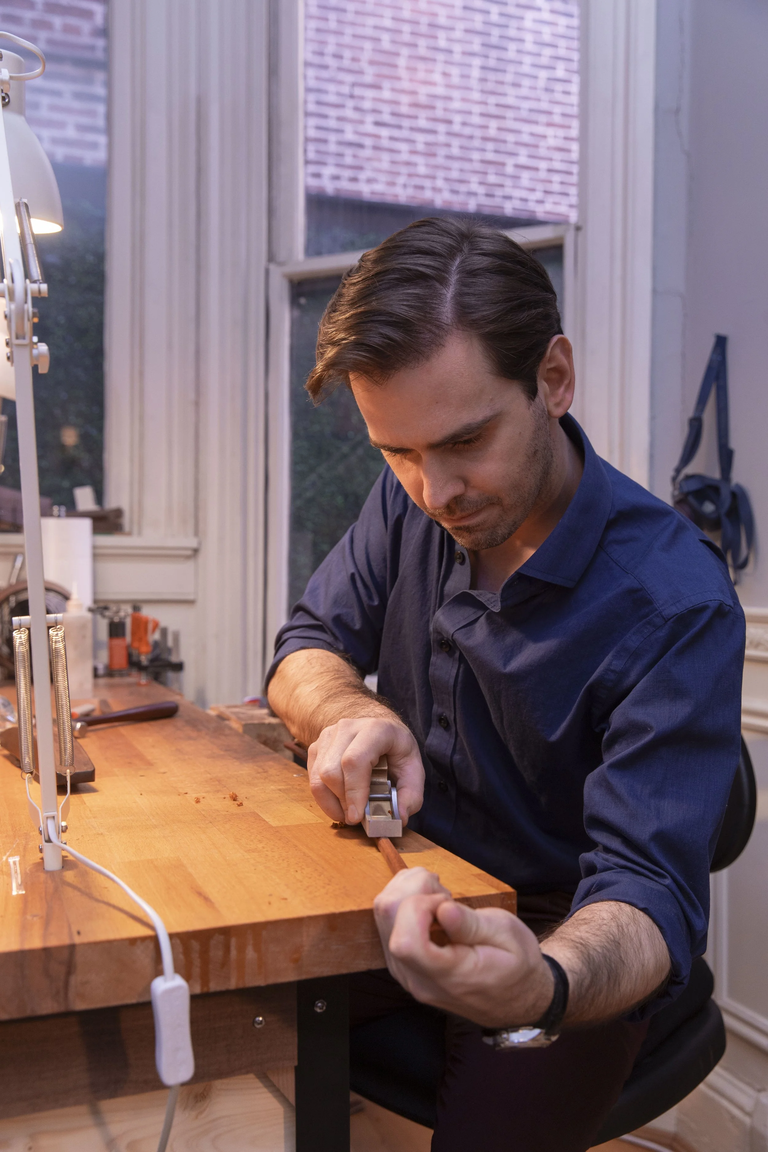 A man woodworking at a workshop table in a well-lit room, using a hand tool on a piece of wood.