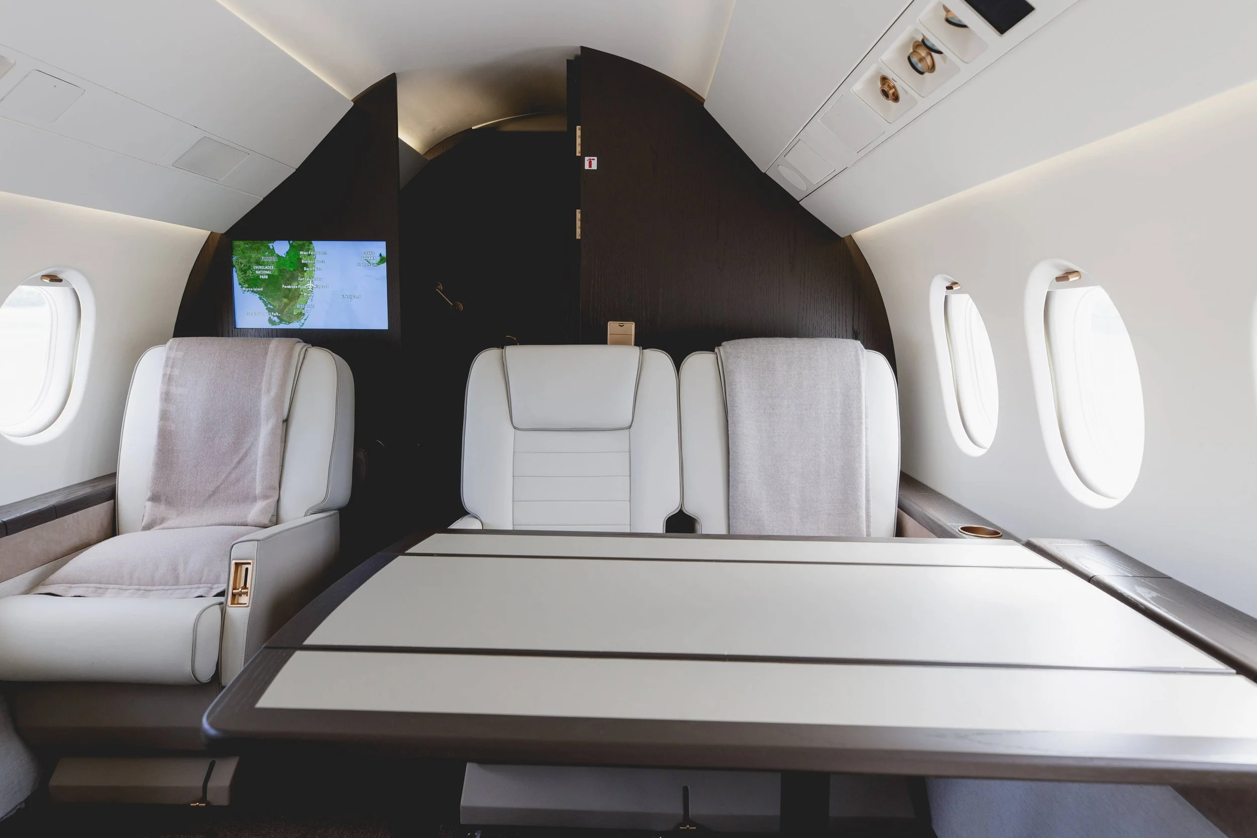 Interior of a private jet with three white leather seats, a fold-out table, small windows, and a screen showing a map of South America.