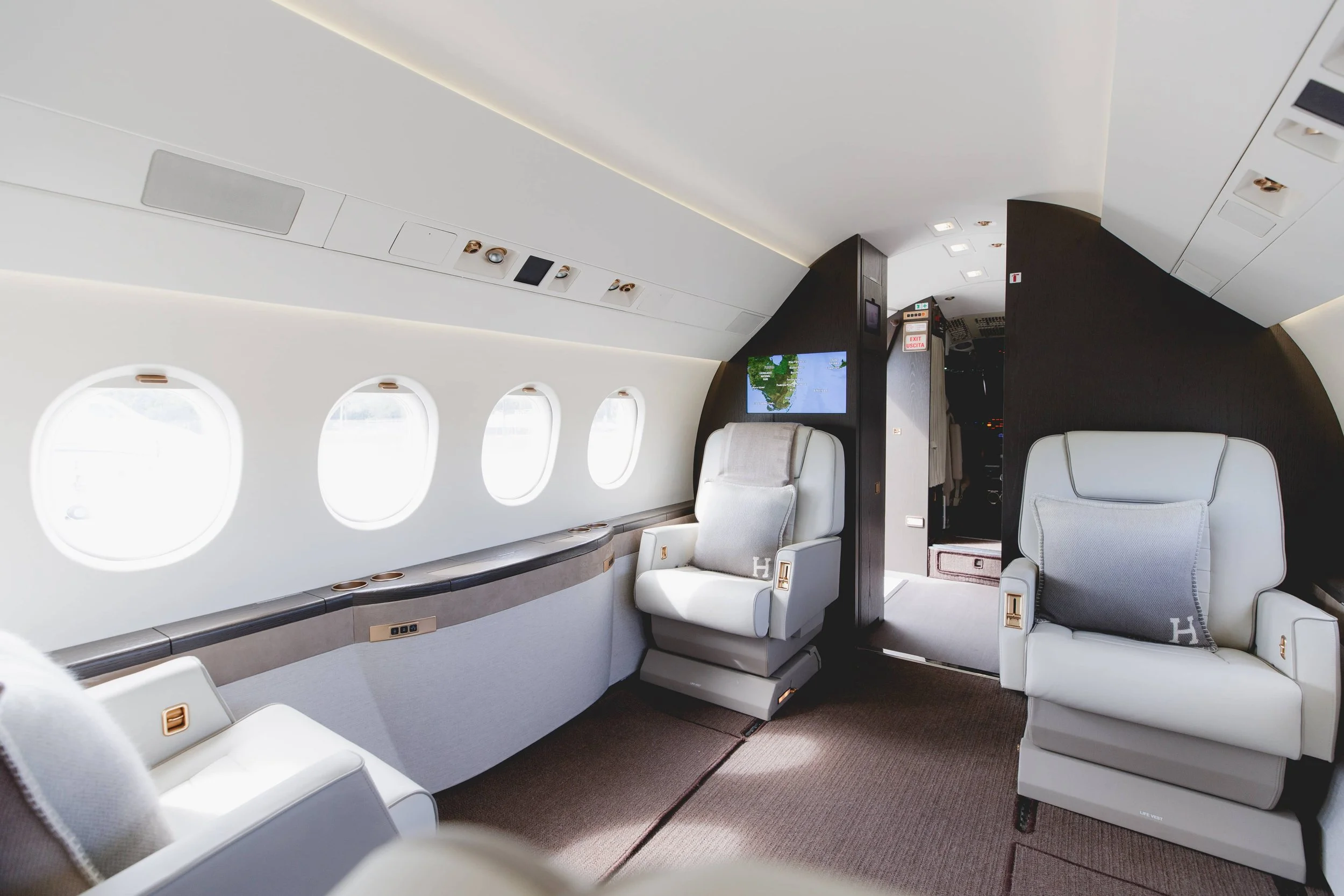 Interior of a luxurious private jet with white leather seats, oval windows, and a small screen showing a map.