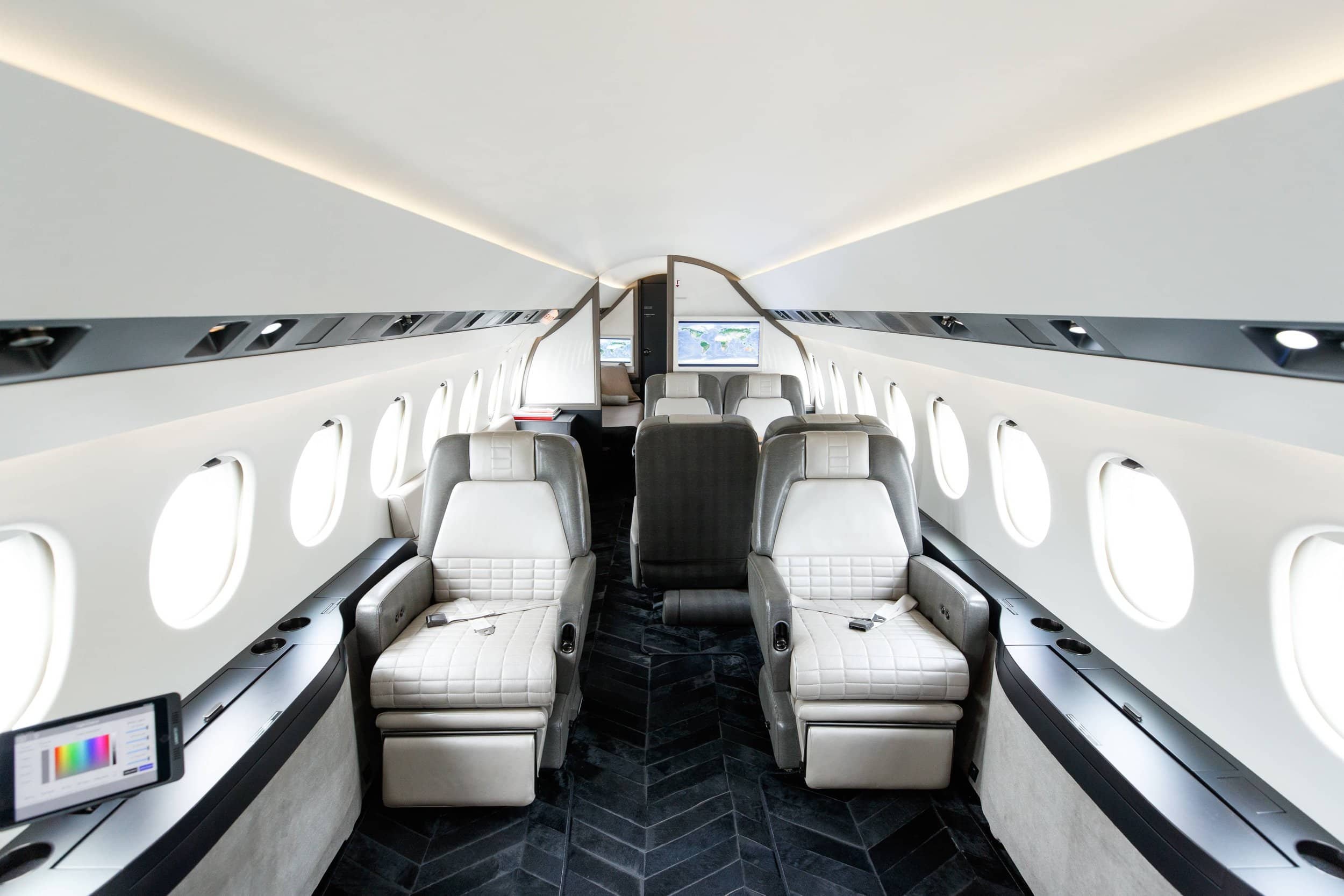 Interior of a private jet with gray and white leather seats, windows along both sides, and a TV screen at the front.
