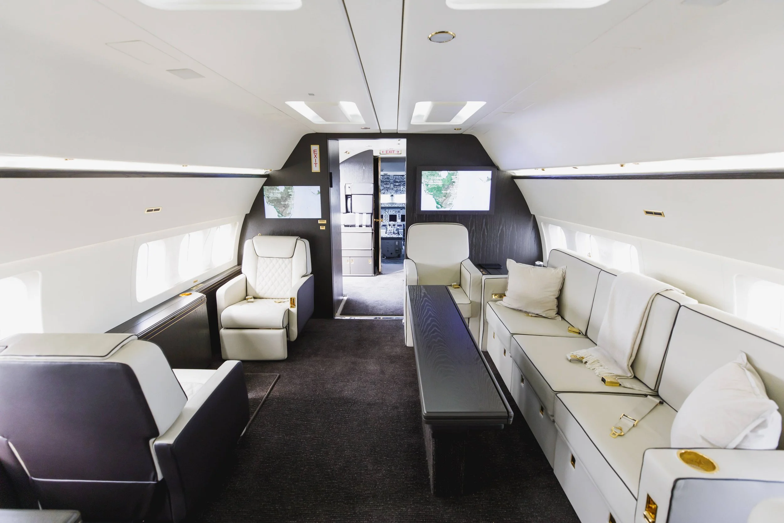 Interior of a private jet with white leather seats, a long black table, and small screens on the walls.