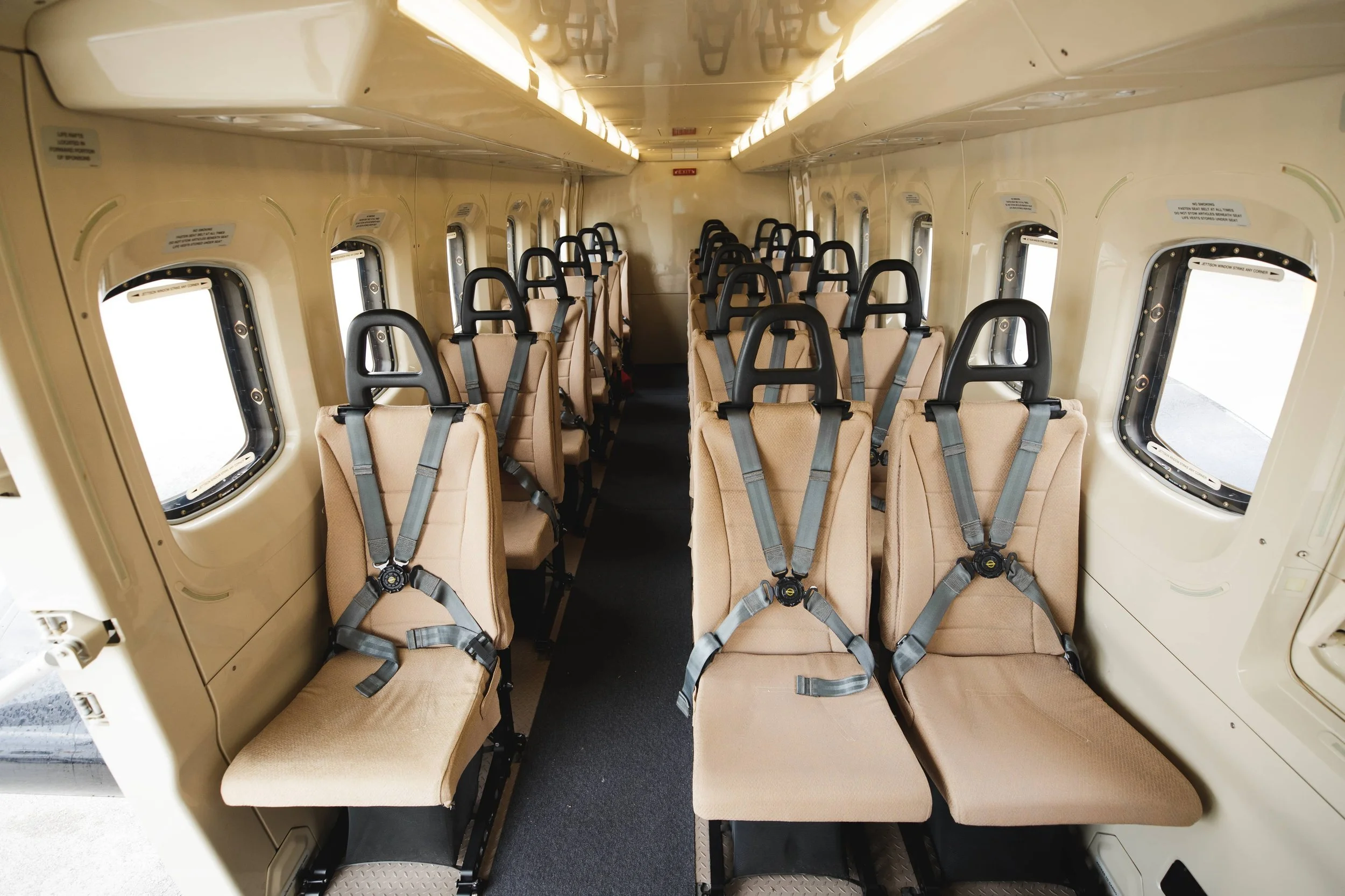 Interior of an airplane with rows of beige seats equipped with seat belts, small oval windows on sides, and a narrow aisle in the middle.