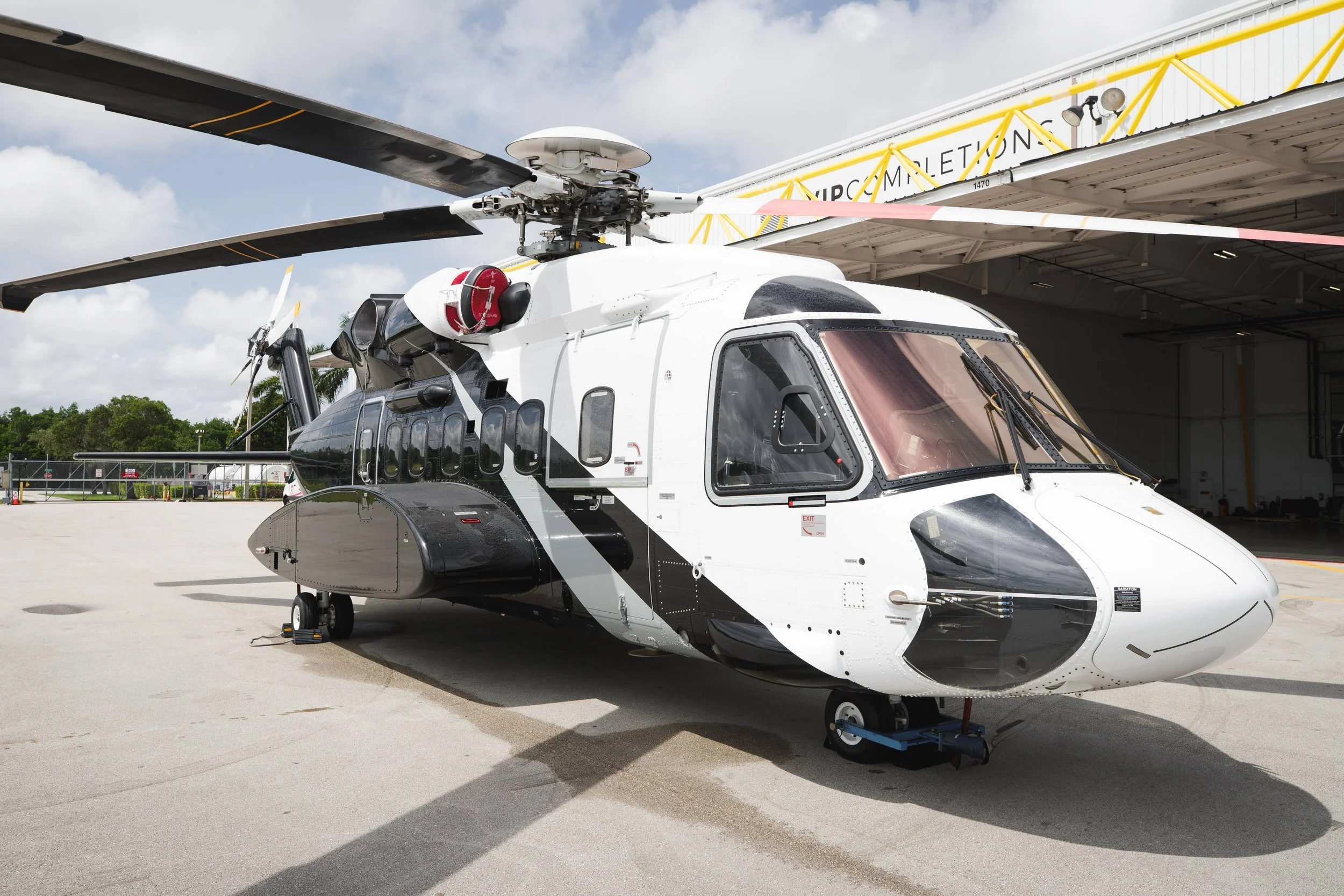 A white and black helicopter parked outside near an aircraft shelter with yellow beams, under a partly cloudy sky.