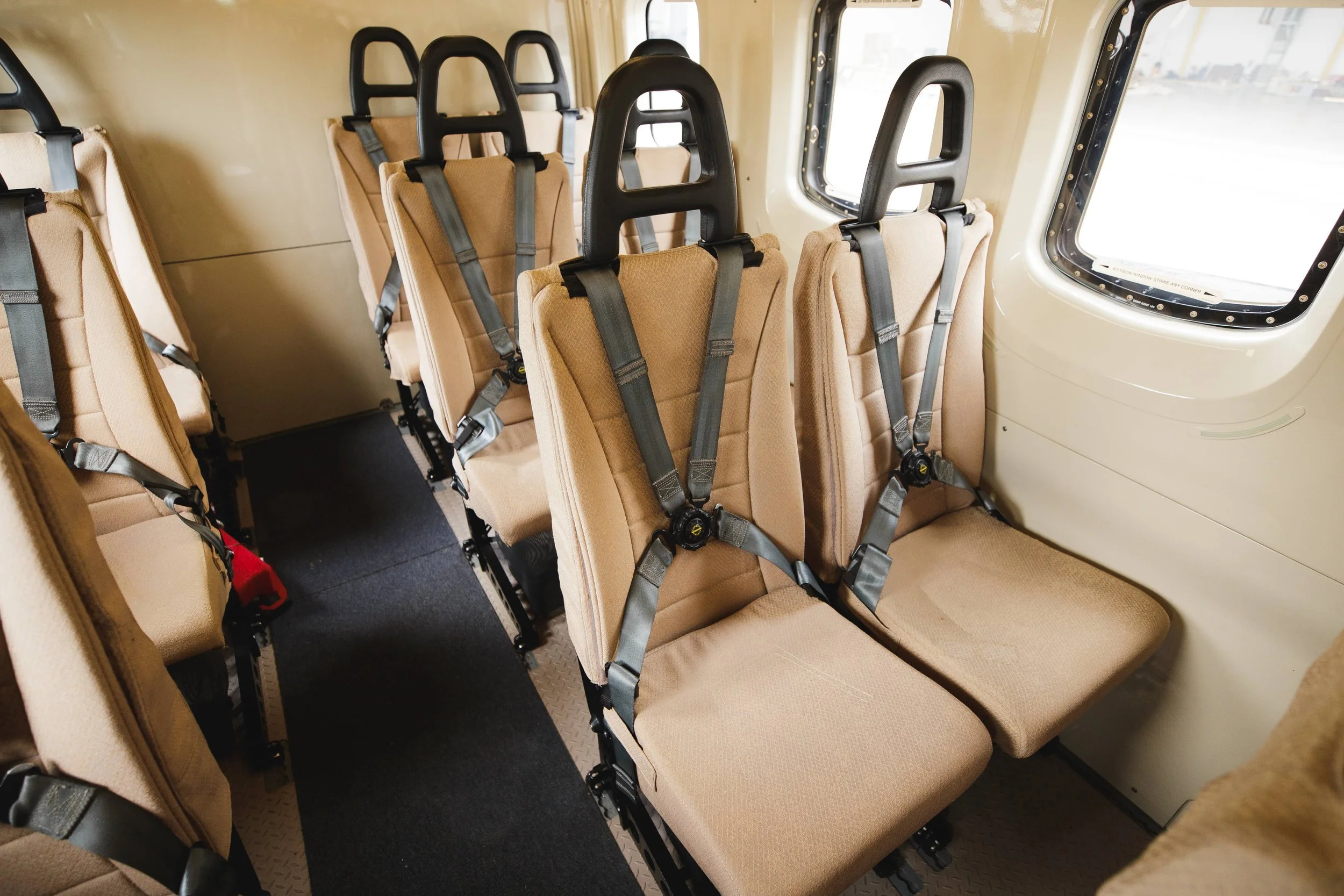Interior of a bus showing beige passenger seats with safety straps, and two windows on the right side.