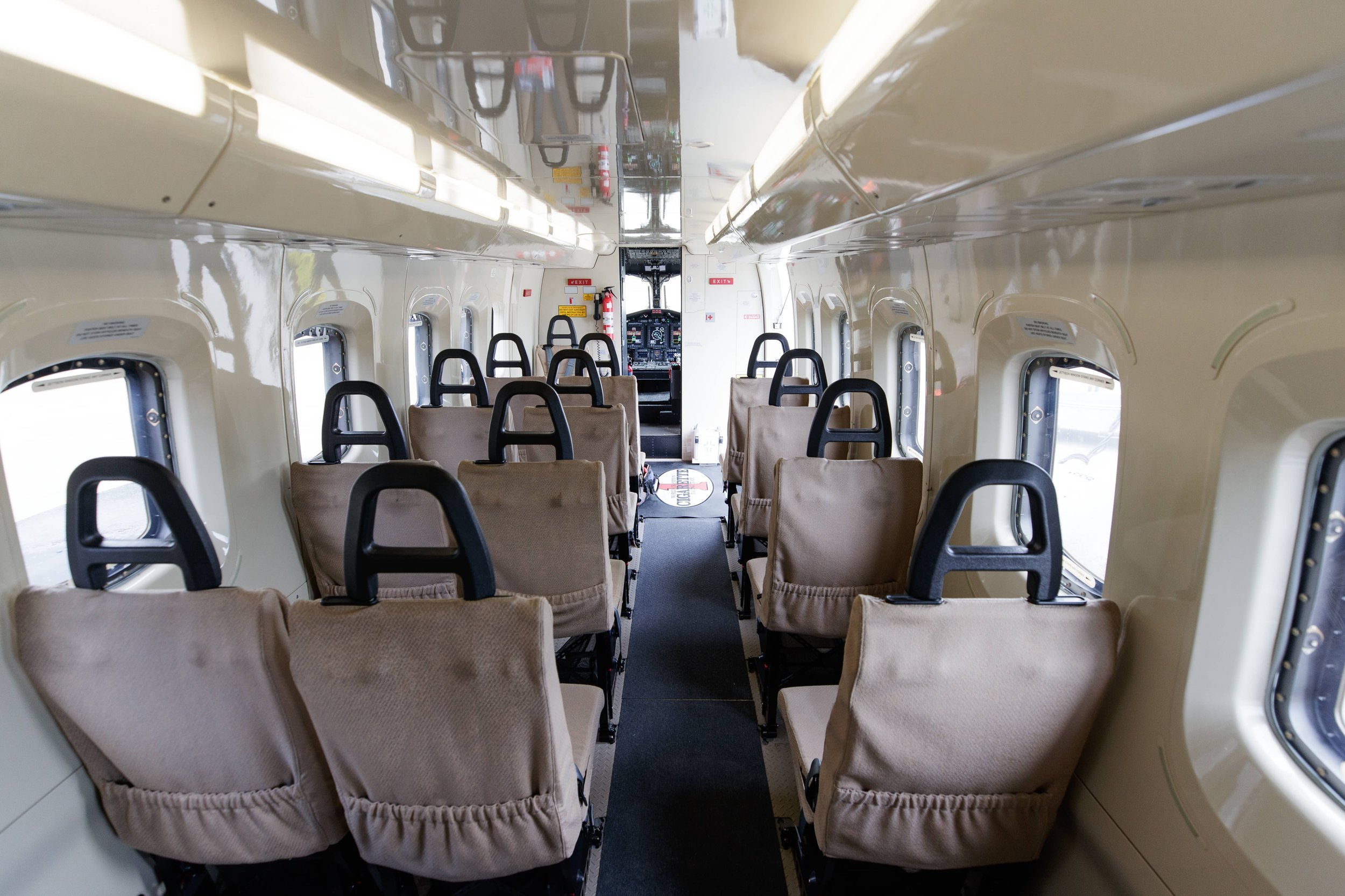 Interior view of a small airplane cabin with multiple beige fabric seats with black handles, windows on both sides, and the cockpit visible at the front.