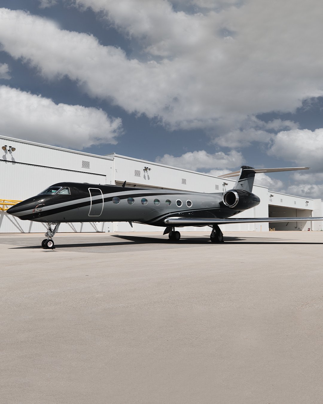 A sleek black private jet parked on an airport tarmac near a large white hangar with cloudy sky in the background.