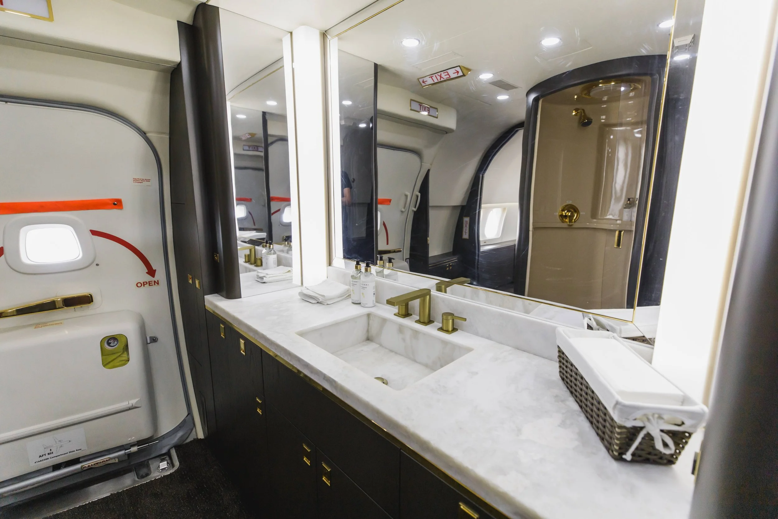 A bathroom vanity with a marble countertop, gold fixtures, a sink, and a large mirror reflecting the restroom area in an airplane.