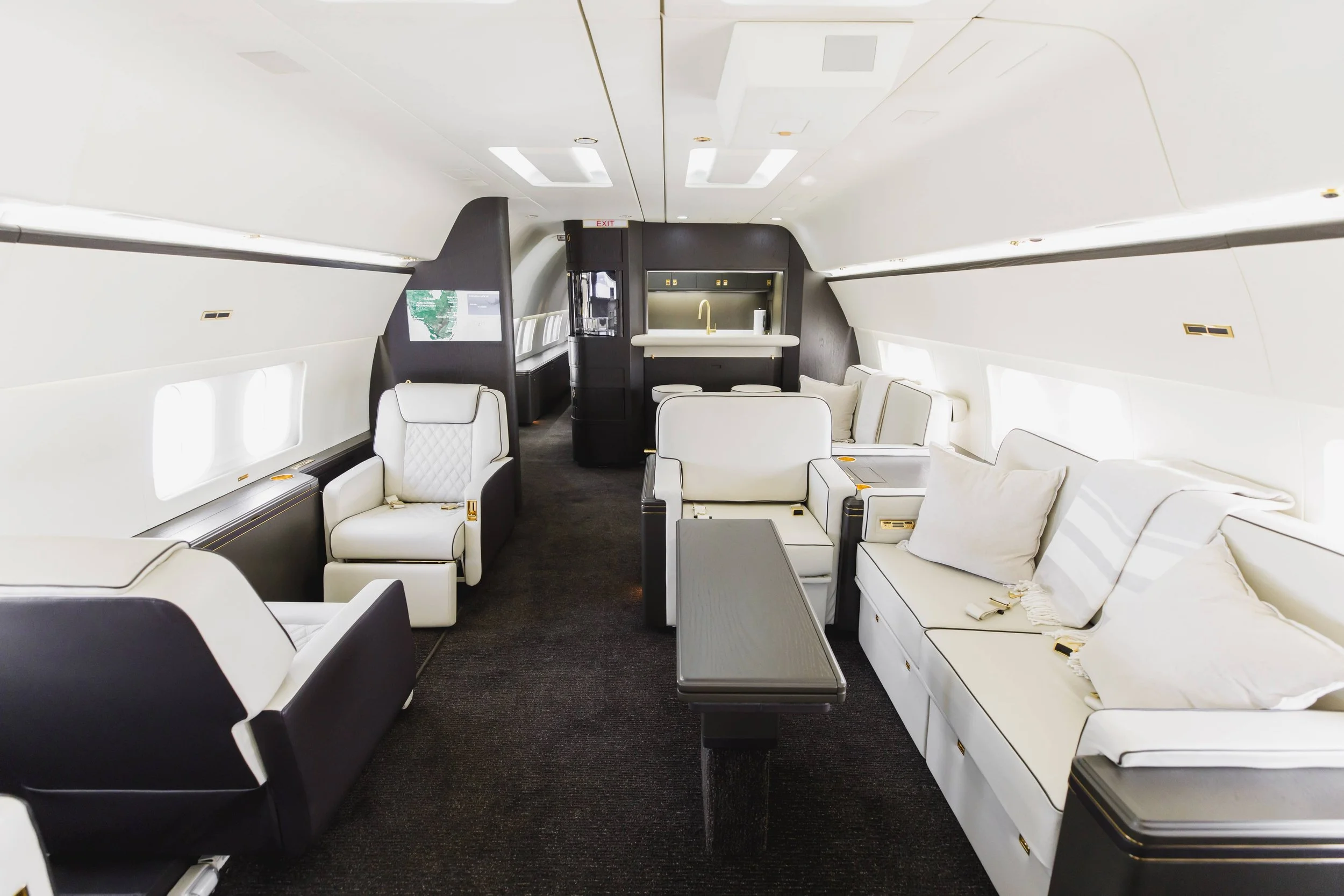 Luxury airplane interior with white and black leather seating, small tables, a kitchenette with a sink, and windows along the sides.
