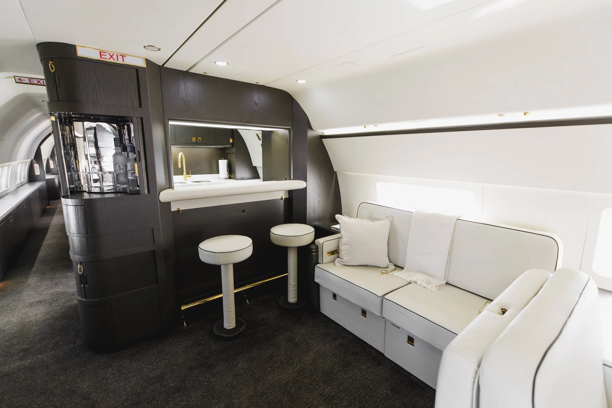 Interior of an airplane lounge with a white sofa, pillows, a bar area with white stools, and a small kitchen in the background.